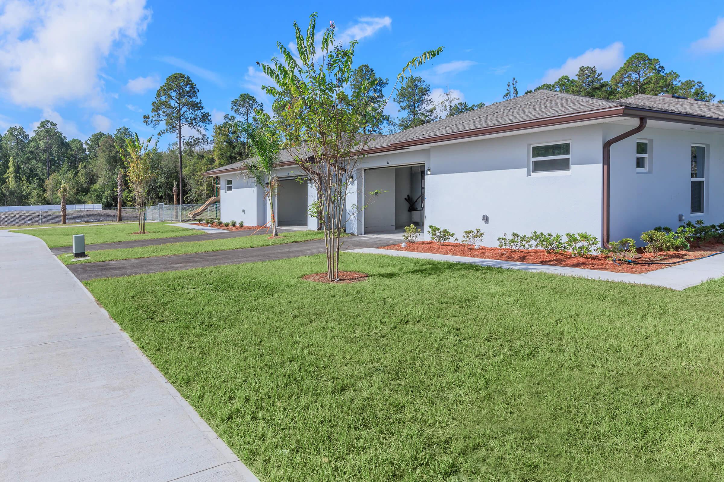 A modern residential building with a light gray exterior, featuring a driveway and landscaped yard. The scene includes manicured grass, small shrubs, and young trees alongside a paved walkway, under a bright blue sky with a few clouds.