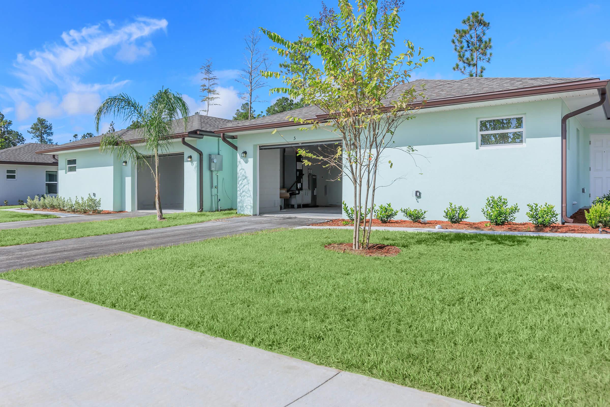 A newly constructed single-story house with a light blue exterior, set in a residential area. The front yard features neatly trimmed grass, small bushes, and a palm tree, while a driveway leads to a garage. The sky is bright blue with a few clouds.