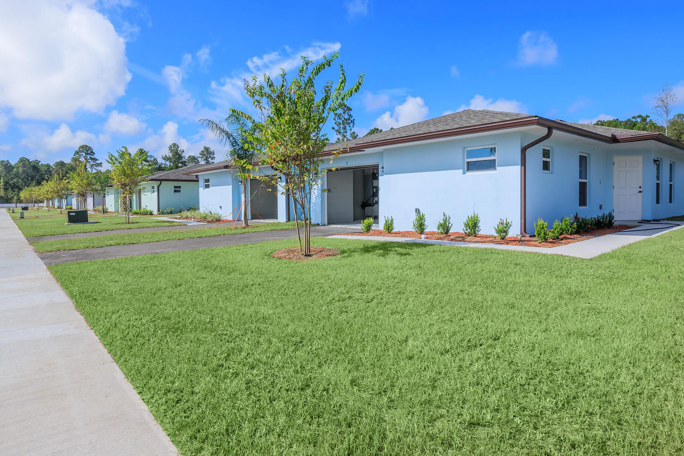 A row of modern, single-story homes with light blue exteriors, surrounded by neatly manicured lawns and small shrubs. The sky is bright blue with a few clouds, and pathways lead to each home, creating a tidy neighborhood appearance.
