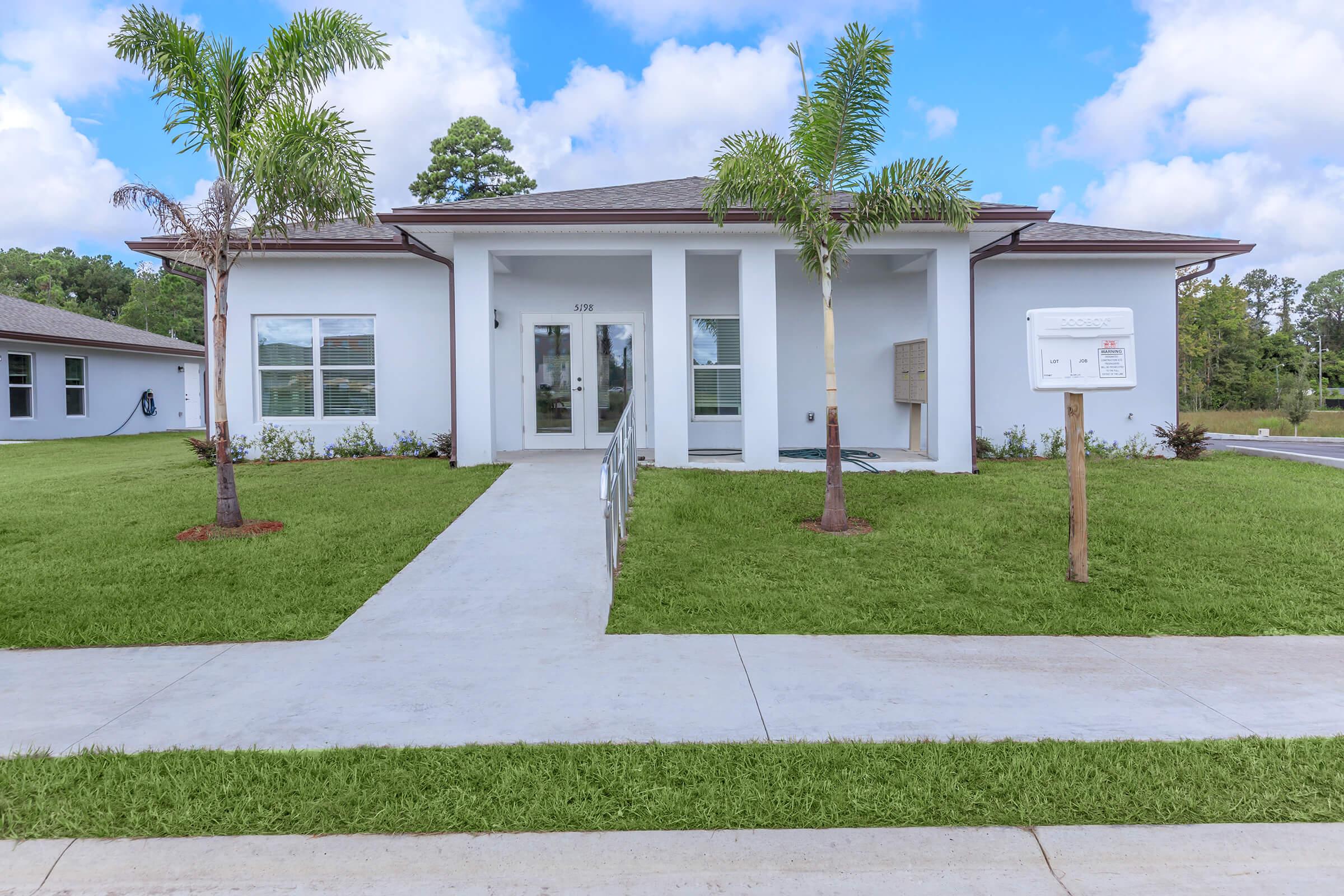 A modern one-story home with a symmetrical facade, featuring a covered front porch and two palm trees on either side. The driveway leads to an accessible entrance, and a mailbox stands on a wooden post near the sidewalk. The surrounding landscape includes well-maintained green grass and a blue sky with clouds.