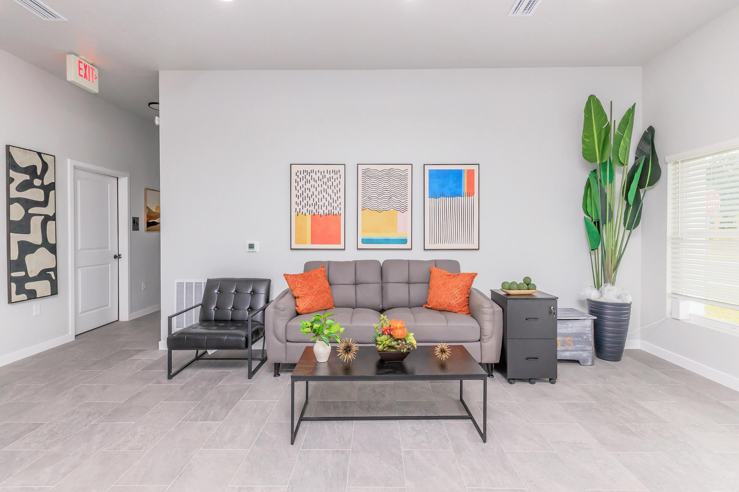 Modern living room featuring a gray sofa with orange accent pillows, a black coffee table, and decorative plants. Artwork in various colors hangs on the wall. The room has light gray walls and floor tiles, with natural light coming through a window.