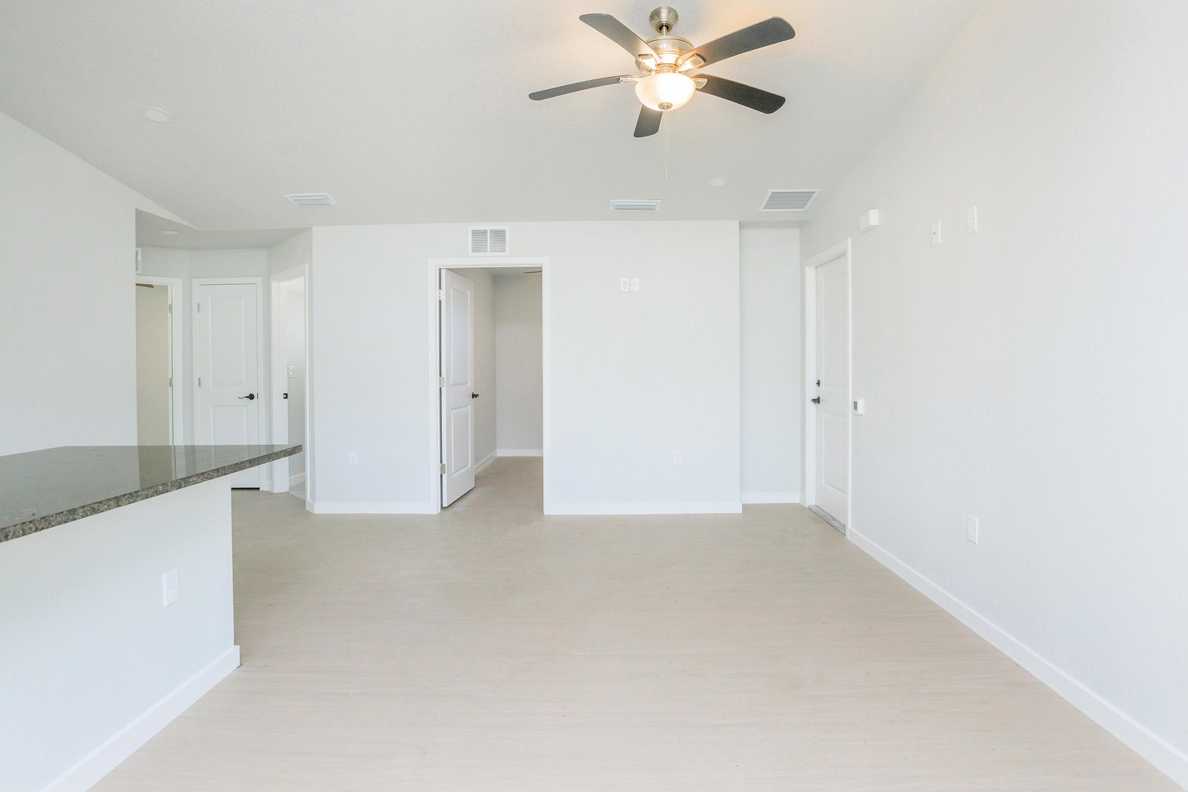 Bright, empty living room with light-colored walls and a smooth beige floor. A ceiling fan is installed in the center, and doorways lead to adjacent rooms. The space is clean and modern, featuring a small kitchen bar area on the left. Natural light fills the room from multiple directions.