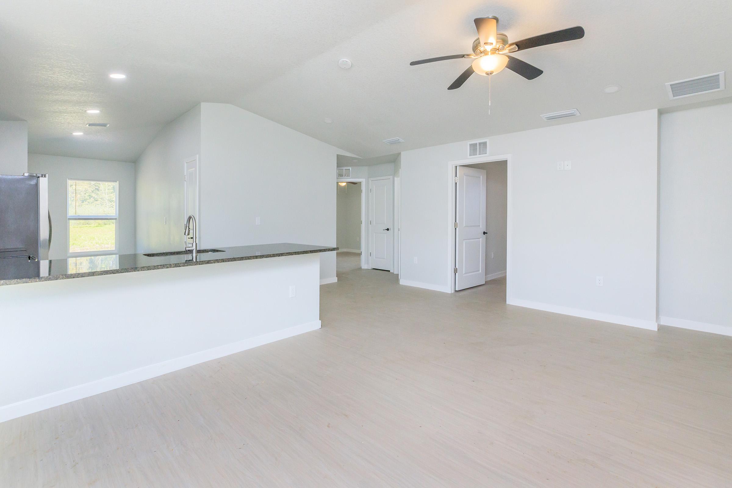 Bright, spacious interior of a modern living area featuring light-colored walls, large windows allowing natural light, and a ceiling fan. The open layout includes a kitchen area with a granite countertop, with doors leading to other rooms, creating a seamless flow in the design.