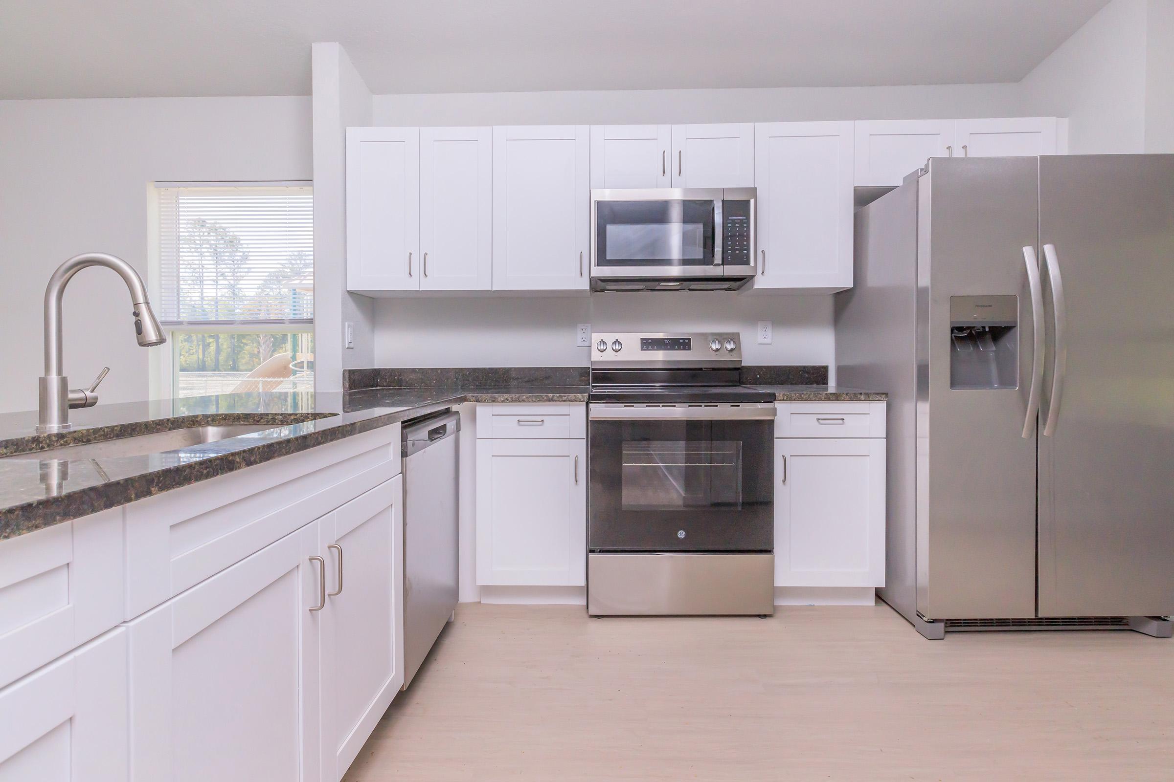 Modern kitchen featuring white cabinetry, a stainless steel refrigerator, a microwave above the stove, an oven, and a sleek sink. The countertops are dark granite, and the space is well-lit by natural light from a nearby window.