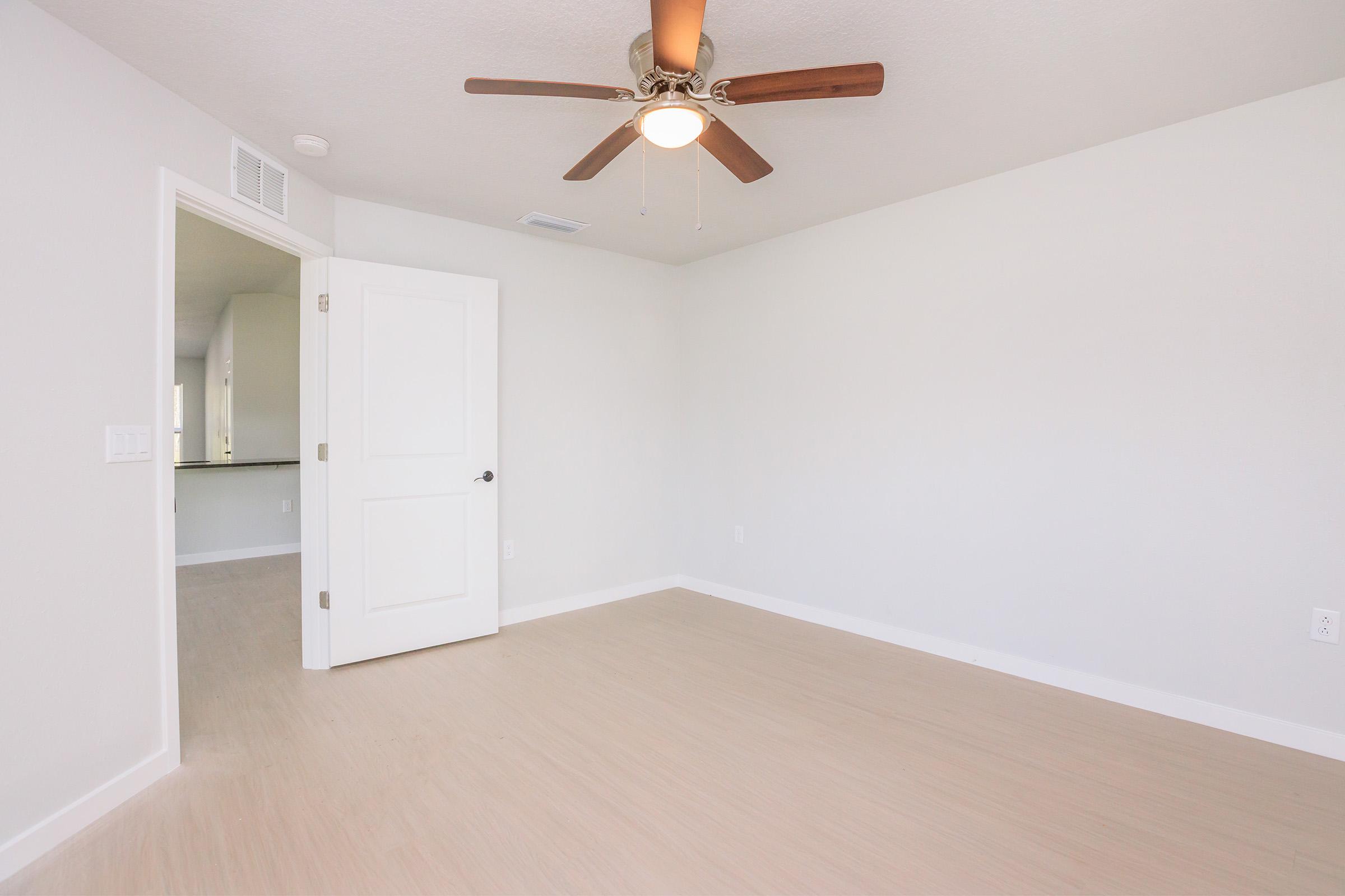 A bright, empty room with light-colored walls and a ceiling fan. The room has a smooth wooden floor and an open door leading to another space. Natural light fills the area, creating a warm and inviting atmosphere.