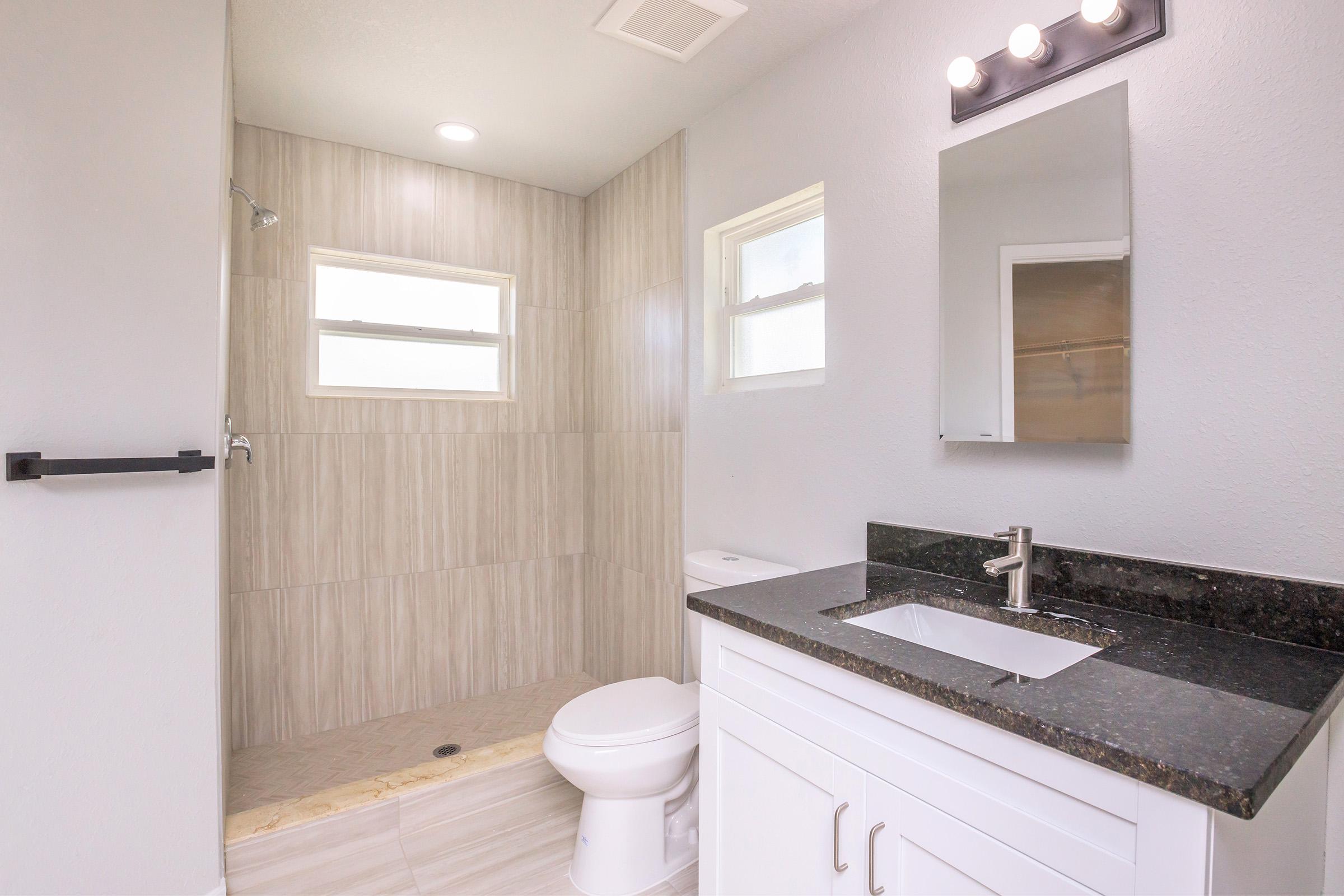 A modern bathroom featuring a walk-in shower with light tiles, a sleek countertop with an under-mount sink, and a white cabinet. The room has grey walls and two windows providing natural light, along with a stylish light fixture above the mirror.