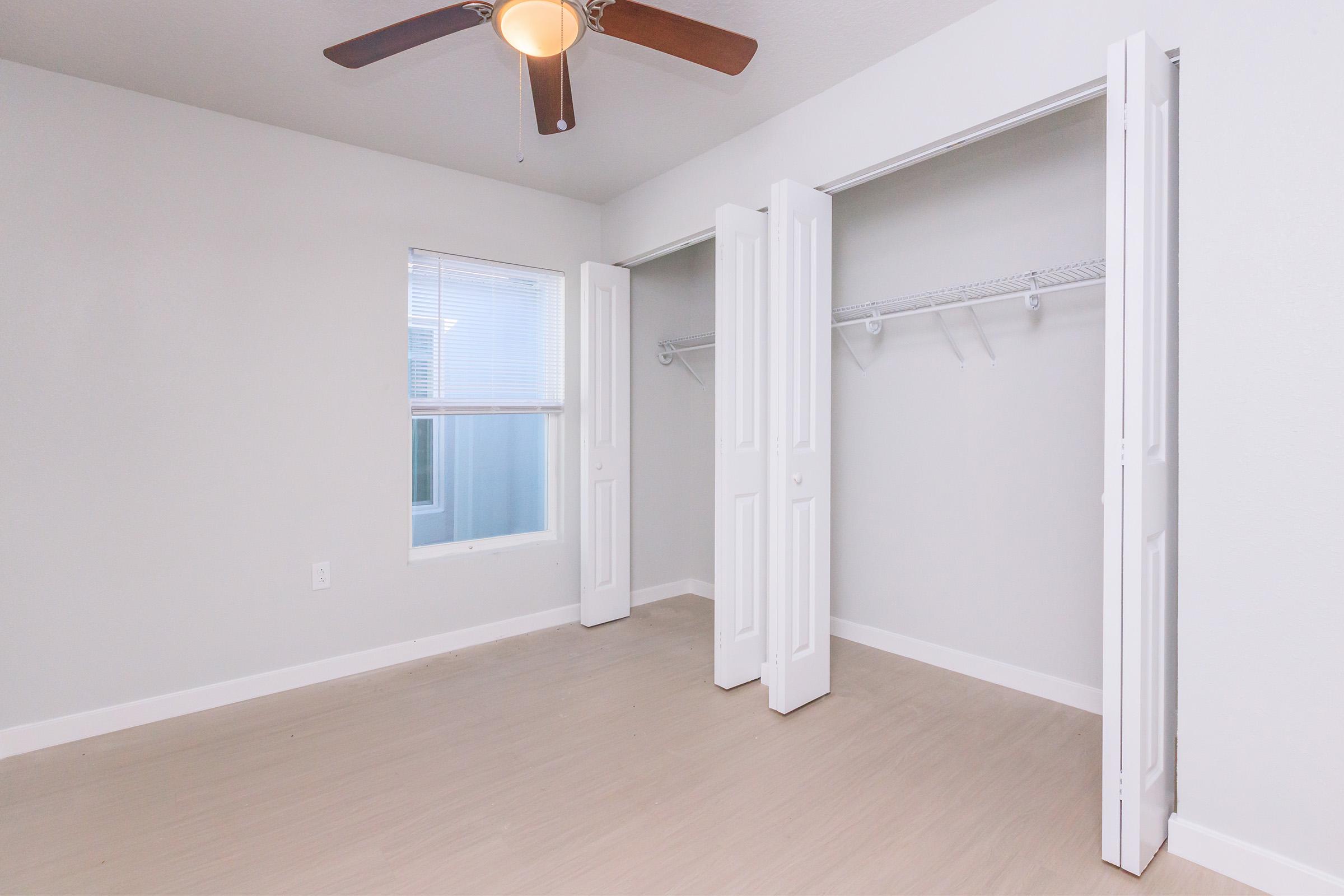 A well-lit, minimalist bedroom featuring light-colored walls and a ceiling fan. There are two open white closet doors with shelving inside. A window allows natural light into the room, which has a light wood or laminate floor.