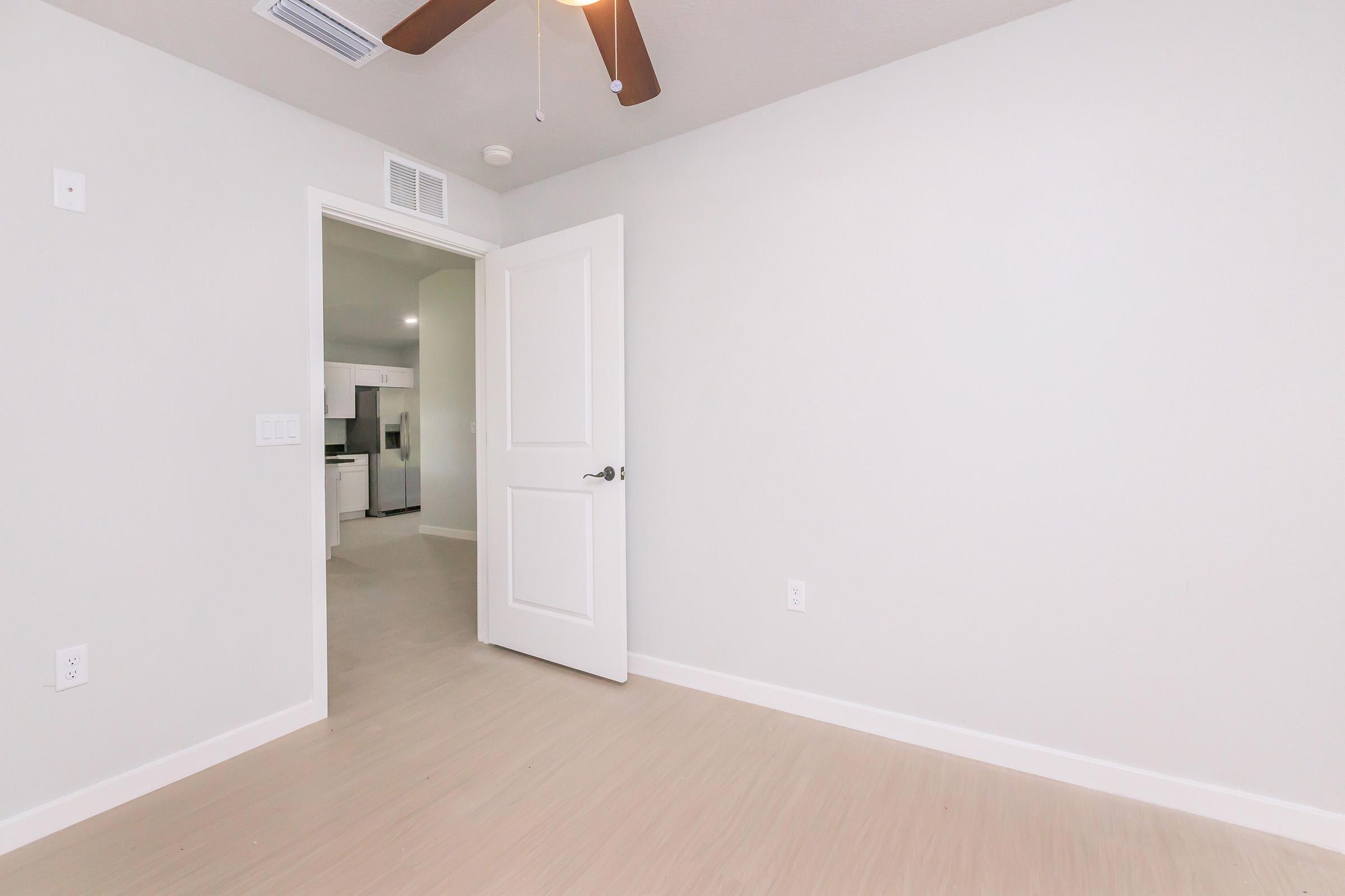 A view of a modern, light-colored room with a closed door leading to another space. The walls are painted in a soft shade, and a ceiling fan is visible. The floor features light-colored wood or laminate. The room is empty, creating a clean and airy atmosphere.