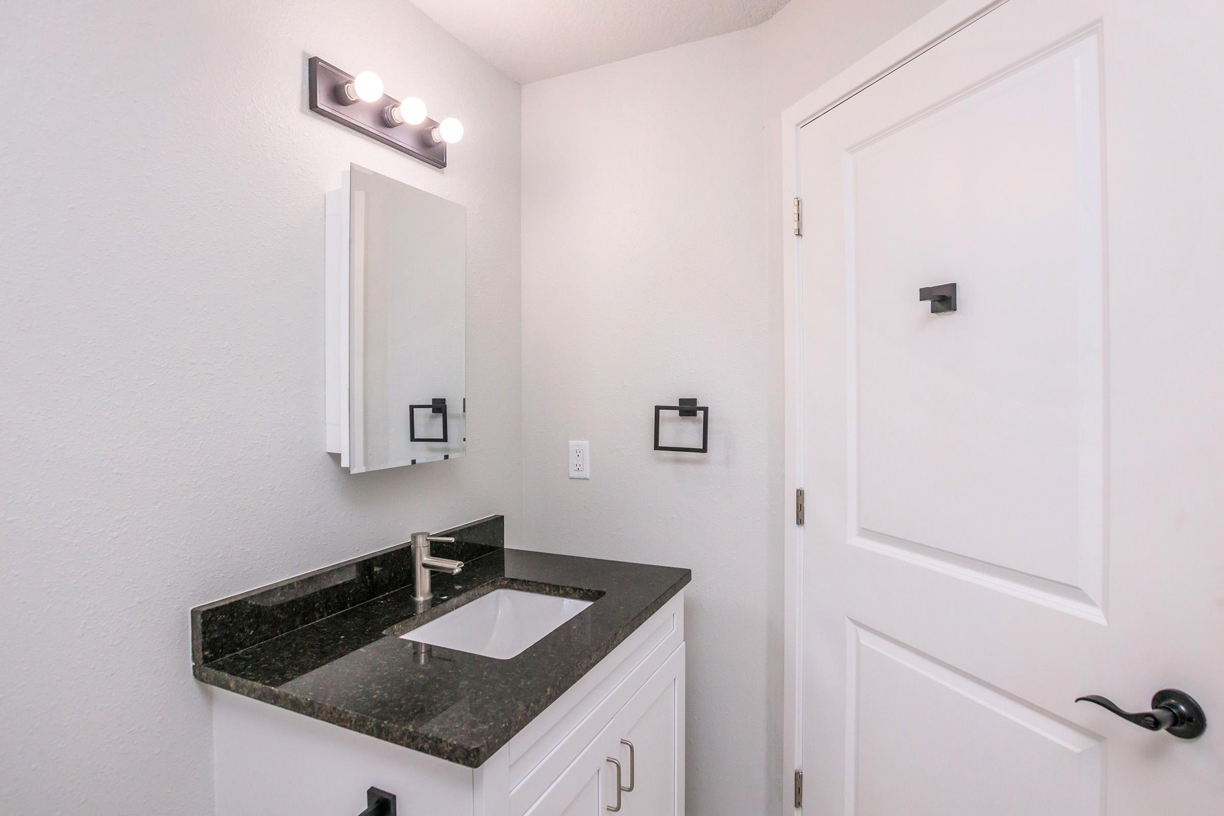 A modern bathroom with light gray walls, featuring a dark countertop and a white sink. There is a mirror above the sink and wall-mounted fixtures for towel storage. The door is white with a simple black handle, and there is overhead lighting with multiple bulbs illuminating the space.