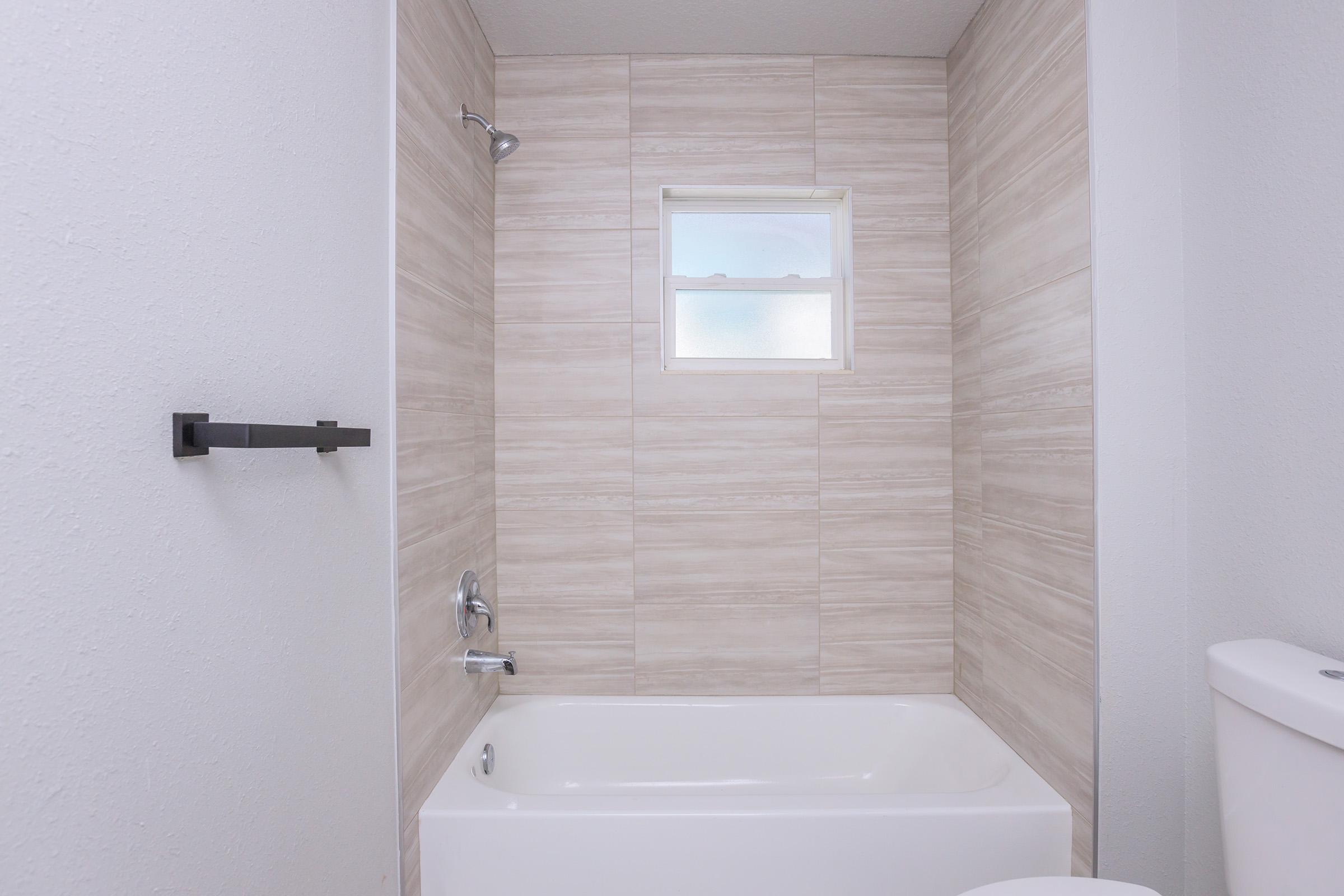 A clean, modern bathroom featuring a bathtub against tiled walls. There is a showerhead above the tub, a small window allowing natural light, and a toilet partially visible on the right. The decor is minimalist with neutral tones, creating a bright and airy atmosphere.