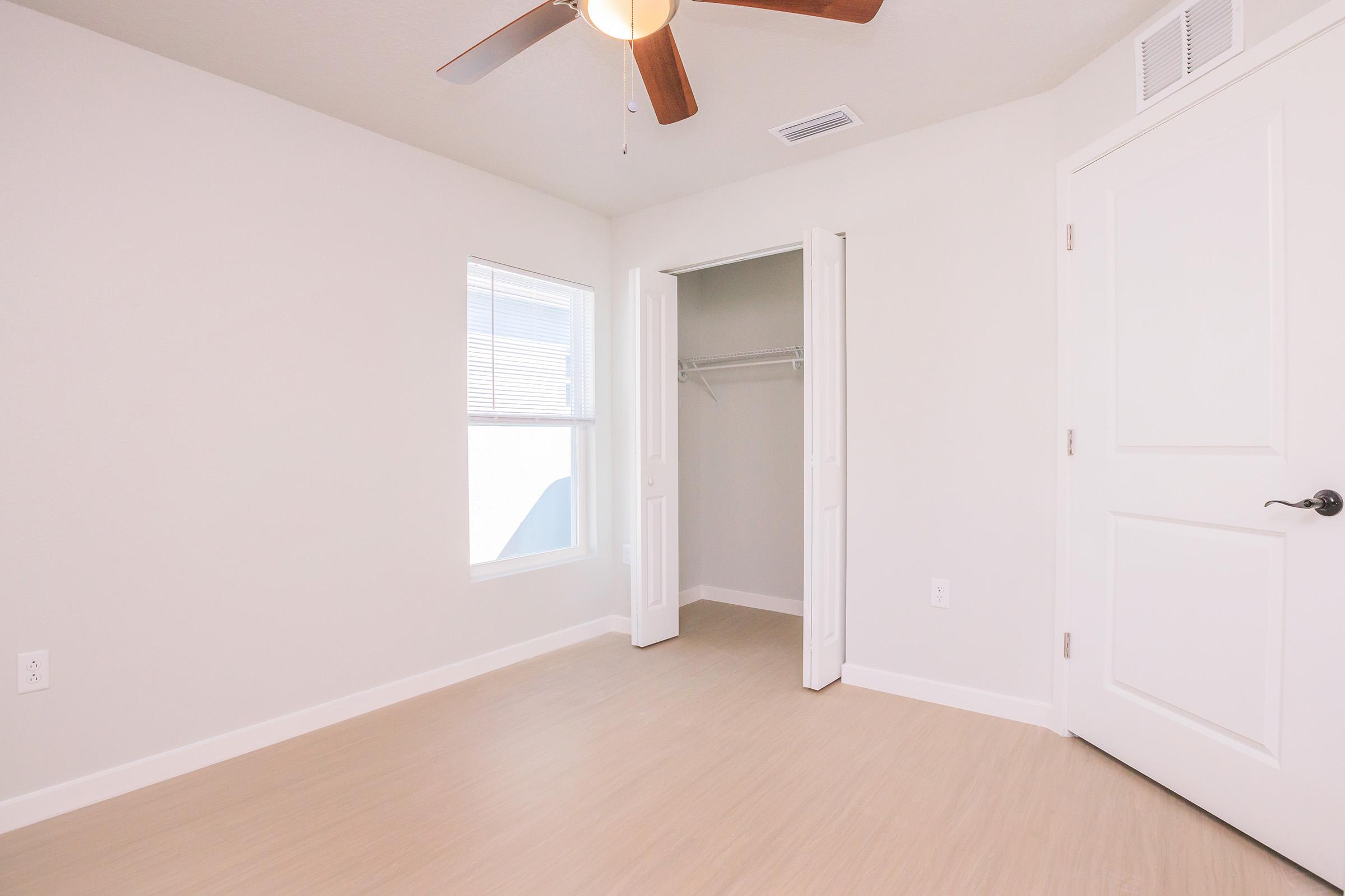 A light and airy bedroom with neutral-colored walls and a wooden ceiling fan. There is a large window letting in natural light and a closet with open double doors. The flooring is a light-colored laminate, creating a spacious and clean appearance.