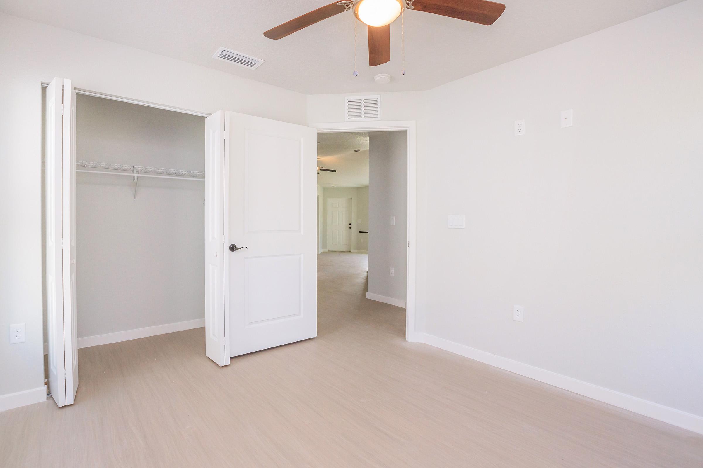 A bright, empty room with light-colored walls and a ceiling fan. One side features an open door leading to a closet with a hanging rod, while the other side has a doorway leading to another room. The flooring is light-colored, creating a spacious and airy atmosphere.