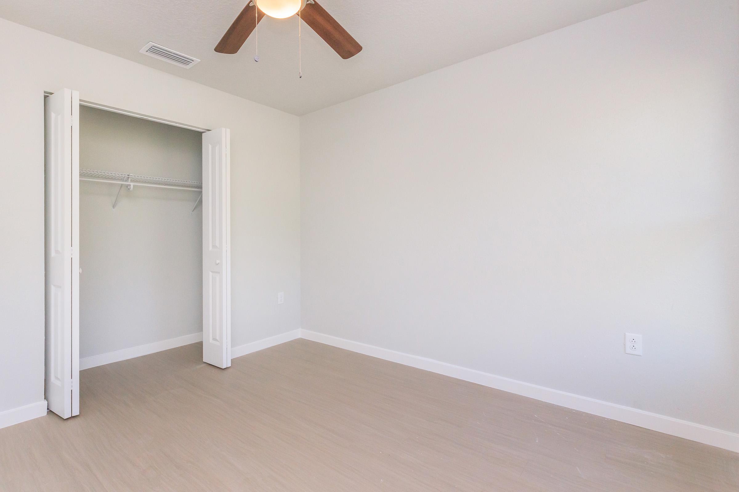 A spacious, empty room with light-colored walls and a ceiling fan. There's a closet with open double doors on the left, revealing a simple shelving unit. The floor has a light wooden texture, creating a bright and airy atmosphere.