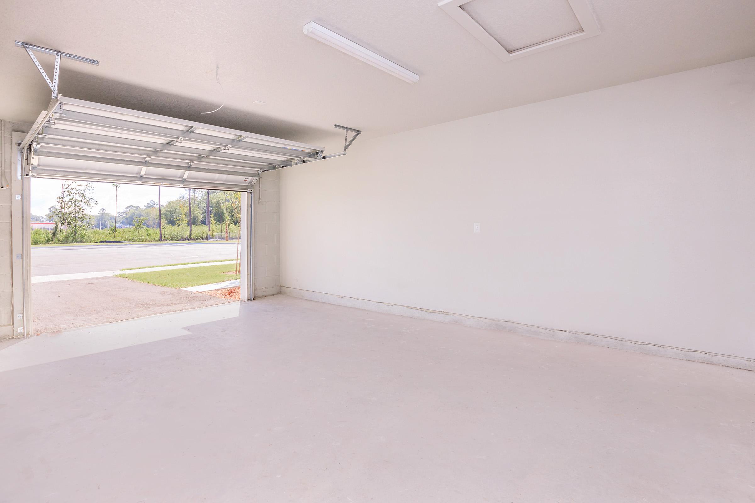 An empty garage with a wide opening showing an outdoor view. The walls are light-colored, and the floor is smooth and clean. Natural light streams in through the open garage door, illuminating the spacious interior.