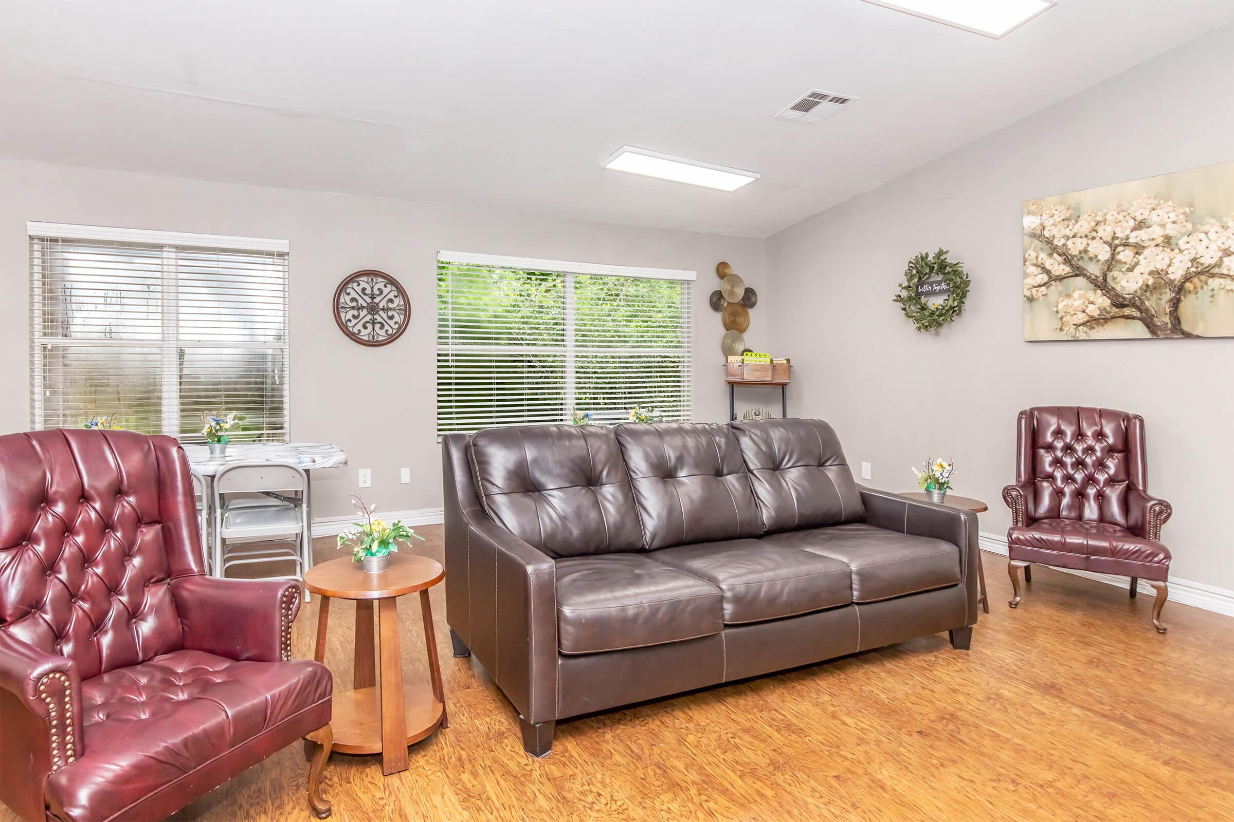 A cozy waiting room featuring a dark leather sofa, two brown armchairs, and a round wooden table with small floral arrangements. The walls are light-colored, with windows allowing natural light, and decorative wall art, including a flower painting and a circular clock.