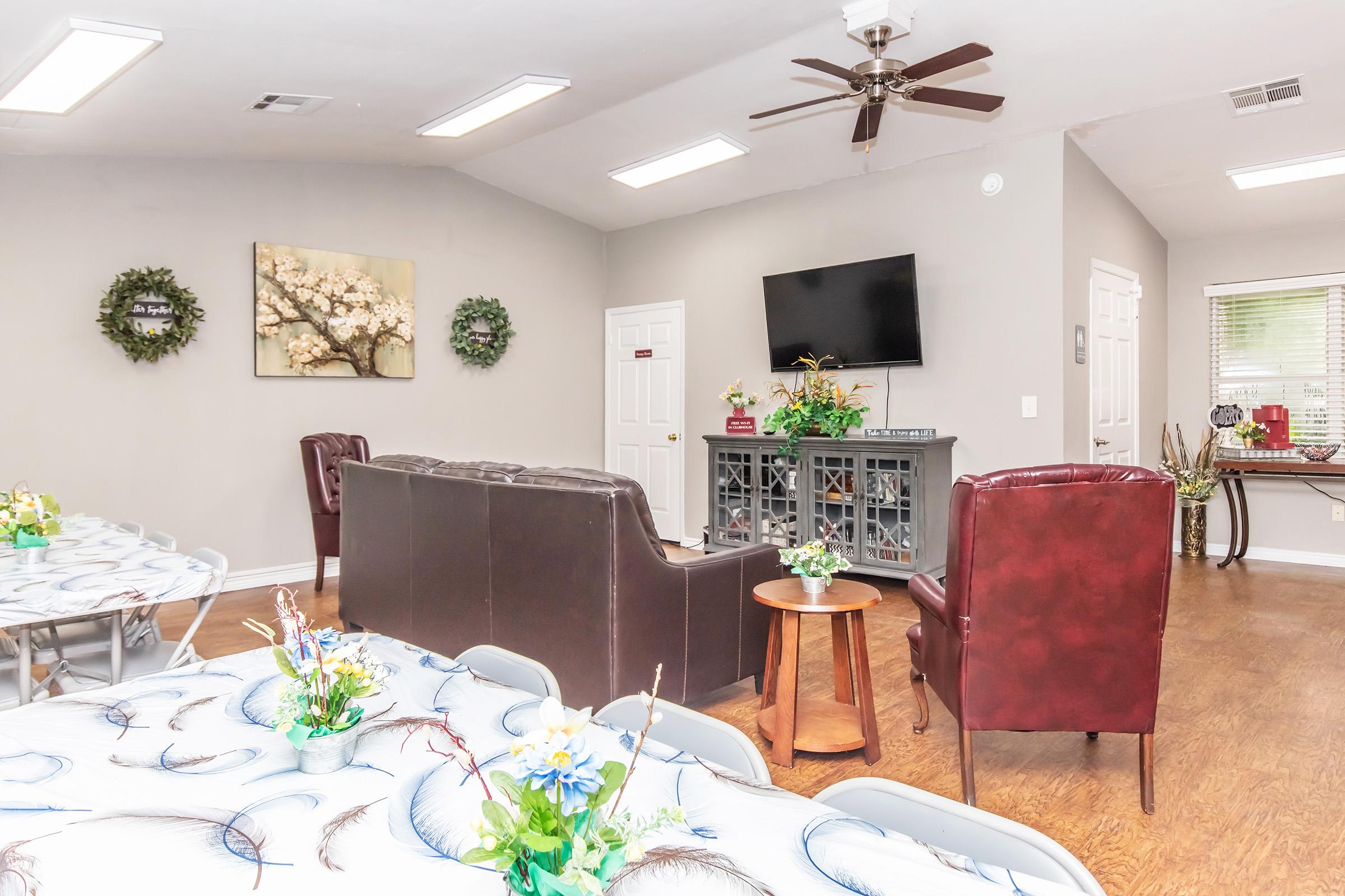 Interior of a cozy room set up for gatherings, featuring a seating area with a dark sofa and burgundy chairs. A wooden table with decorative flowers sits nearby. The walls are adorned with greenery and artwork, and there is a television mounted on the wall. Large windows provide natural light.
