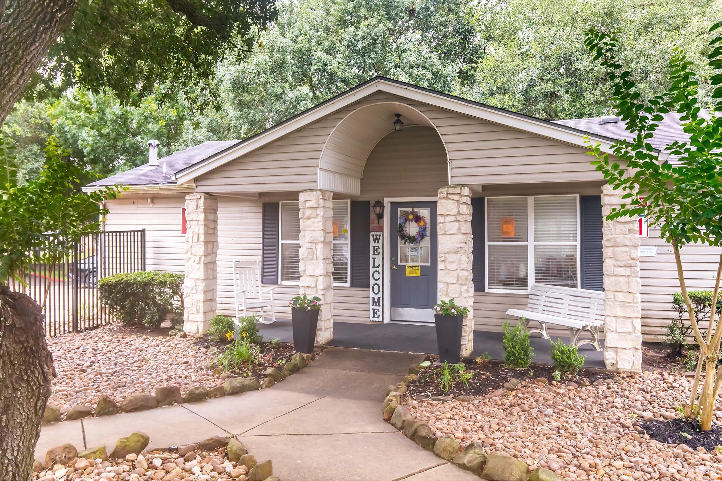 A welcoming entrance of a single-story house with a stone facade, featuring a large archway and a "WELCOME" sign. Two white benches are placed outside, and there are planters with greenery flanking the entrance. The path is lined with small rocks, leading to the door, surrounded by trees and bushes.