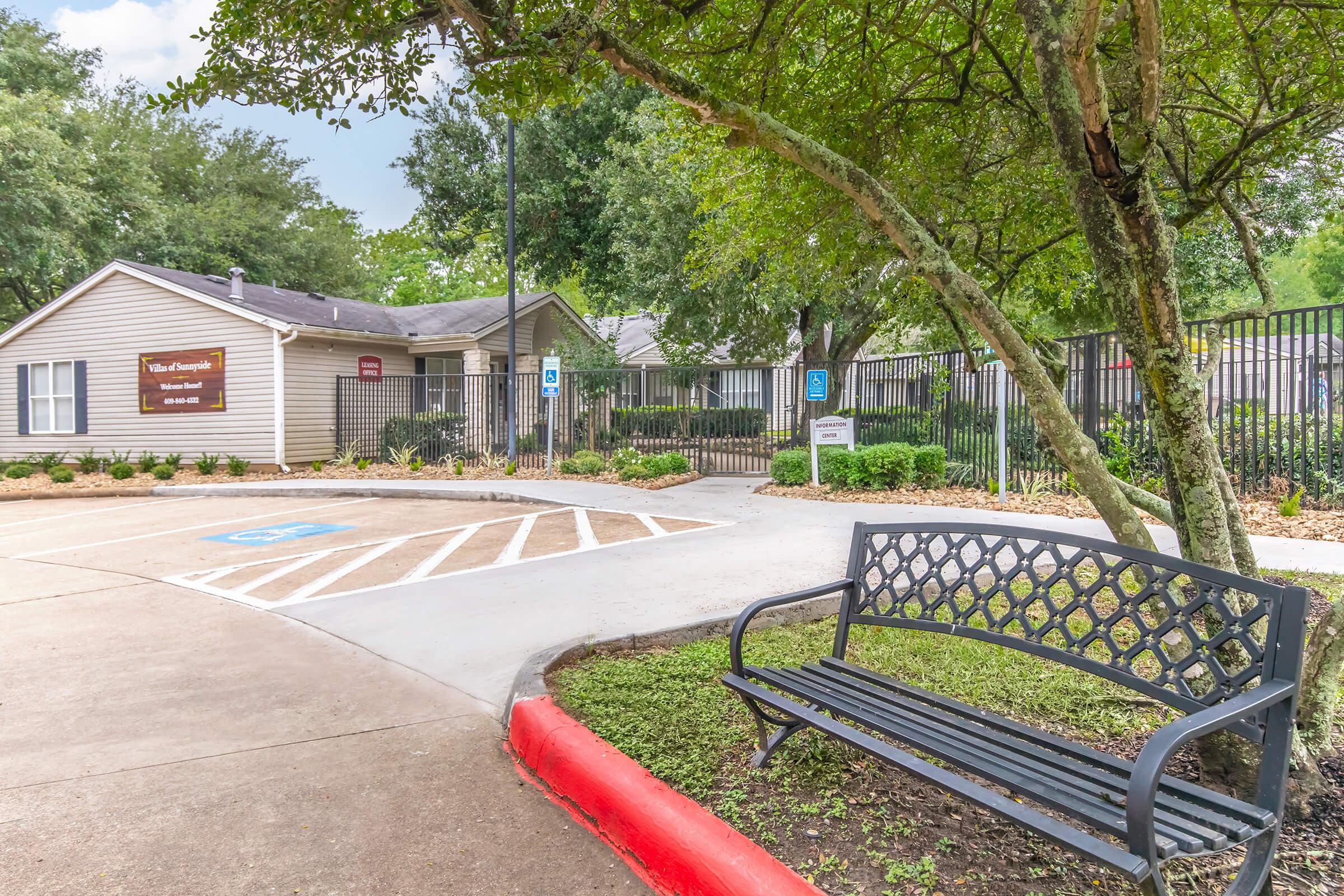 A peaceful neighborhood entrance featuring a well-maintained pathway and a decorative bench under a tree. A sign indicates the community name, and a gated area is visible in the background, surrounded by lush greenery and landscaping. Accessible parking spaces are marked nearby.