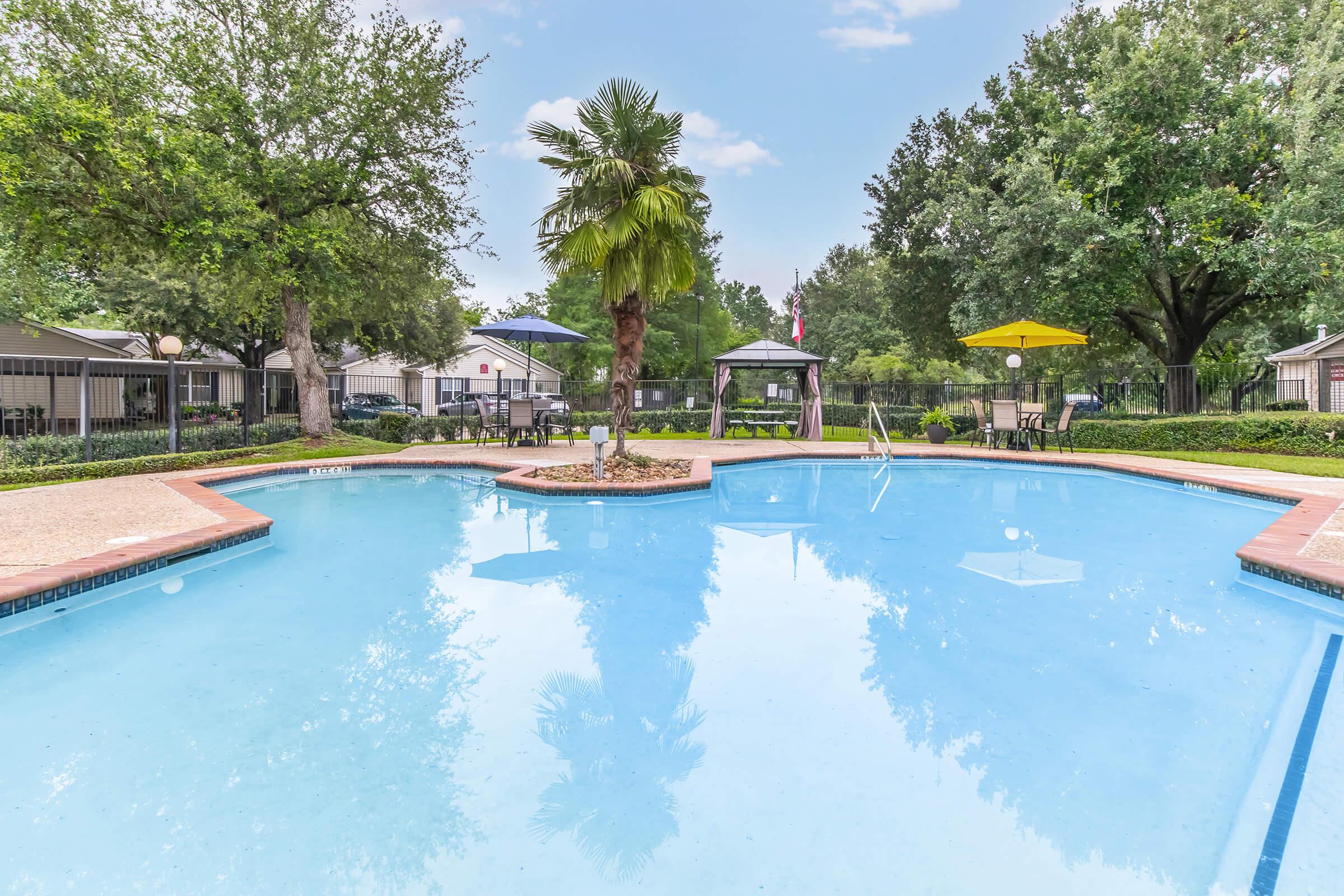 A swimming pool surrounded by lush green trees and a gazebo. In the background, there are umbrellas in yellow and blue, and lounge chairs are set up near the pool. The sky is clear with a few clouds, creating a relaxing outdoor atmosphere.