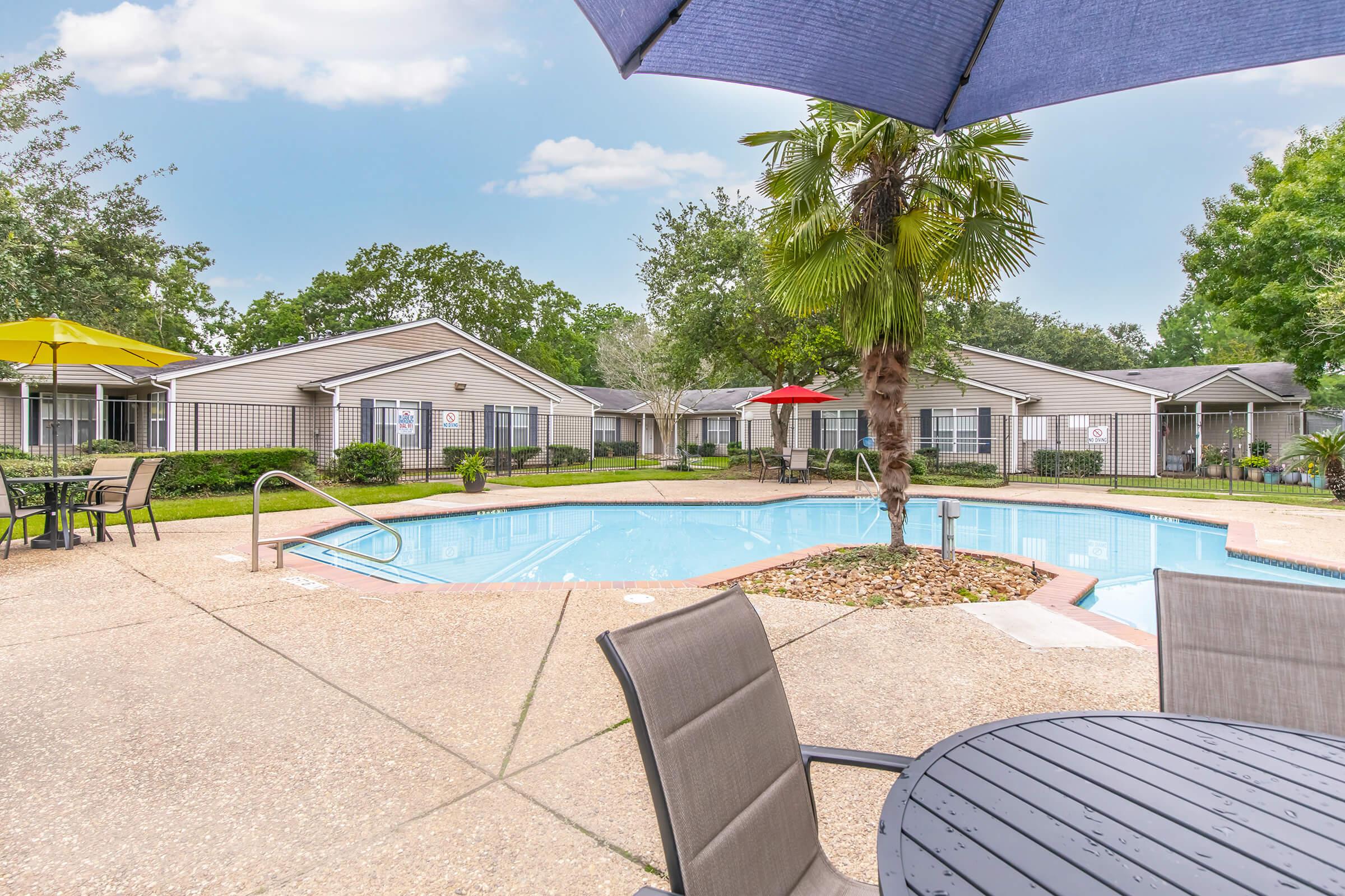 A sparkling blue pool surrounded by lounge chairs, with umbrellas providing shade. In the background, several light-colored buildings are visible, set among lush greenery, including palm trees. The scene is bright and inviting, ideal for relaxation and leisure.