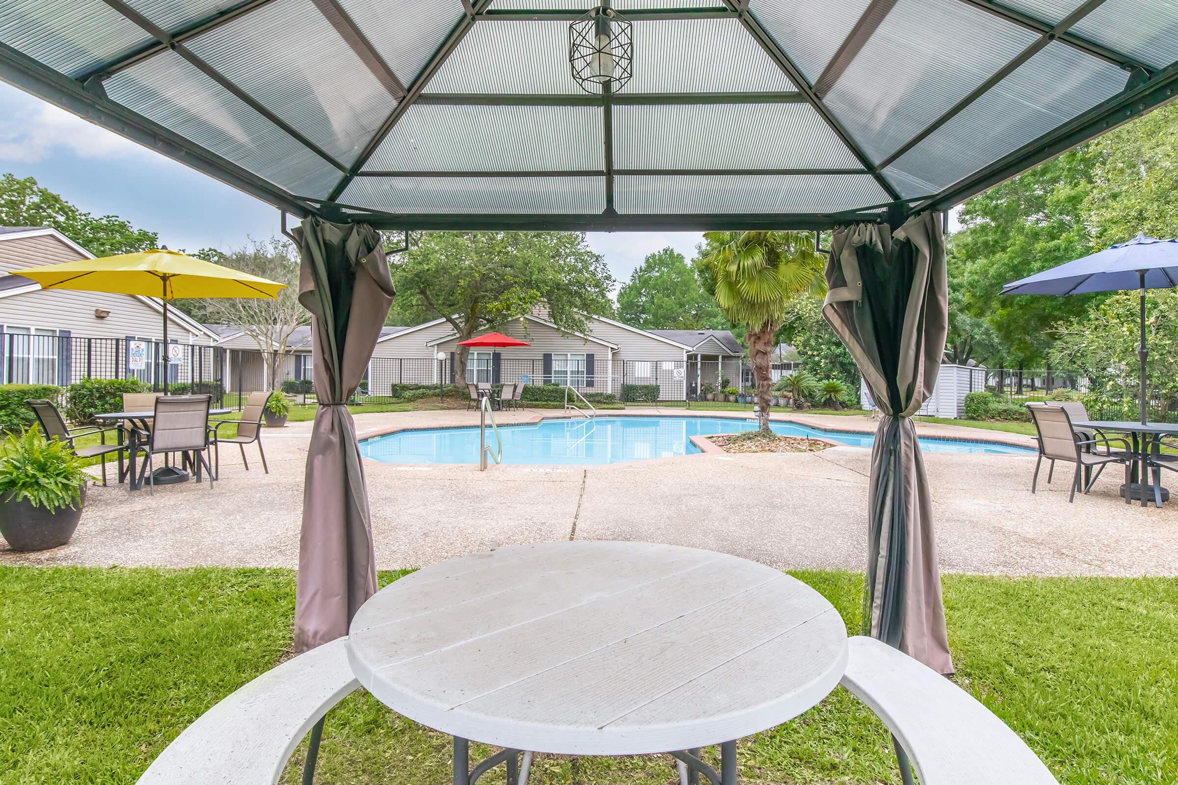 View from a gazebo with a round table overlooking a swimming pool surrounded by green grass and lounge chairs. Colorful umbrellas provide shade, and nearby trees add to the serene atmosphere in a residential setting. The pool area is inviting, with a clean and well-maintained environment.