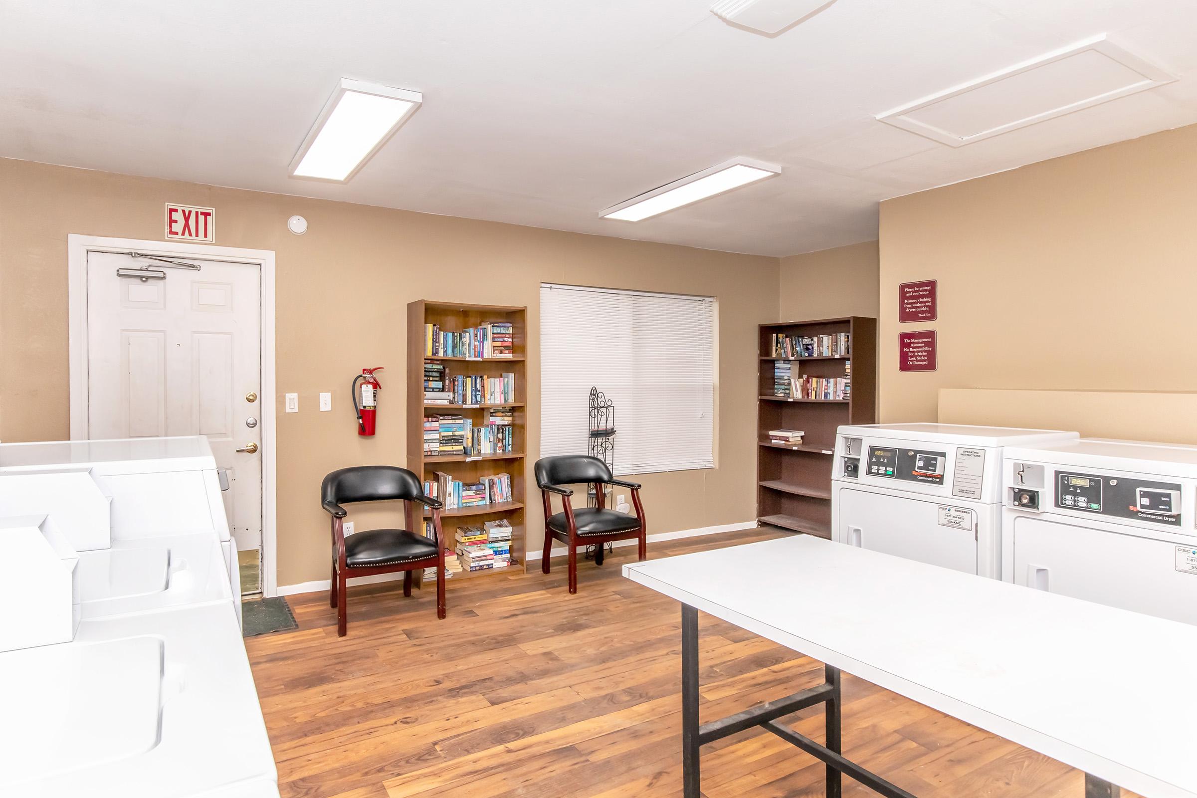 A clean and well-lit laundry room featuring several washing machines and dryers. There are two chairs for seating and a bookshelf filled with books. A window provides natural light, and there's an exit door visible in the background. The walls are painted in a warm beige color, contributing to a cozy atmosphere.