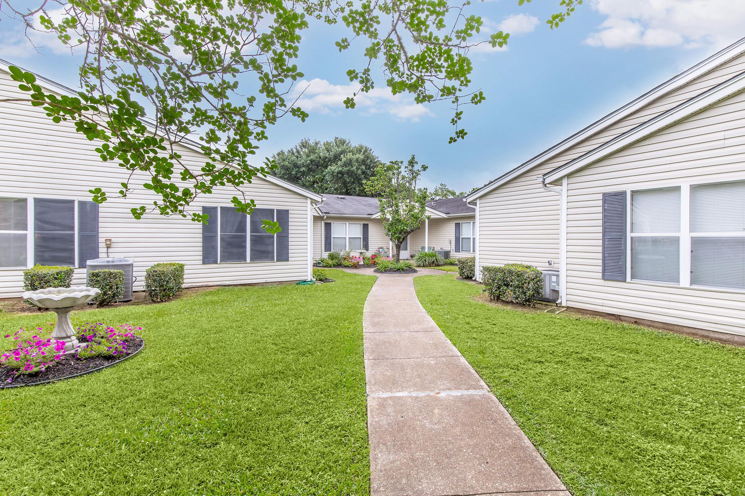 A well-maintained pathway leads through a green lawn surrounded by single-story residential buildings. Flowerbeds with vibrant flowers and neatly trimmed bushes flank the path. The sky is bright with a few clouds, creating a serene and inviting atmosphere.