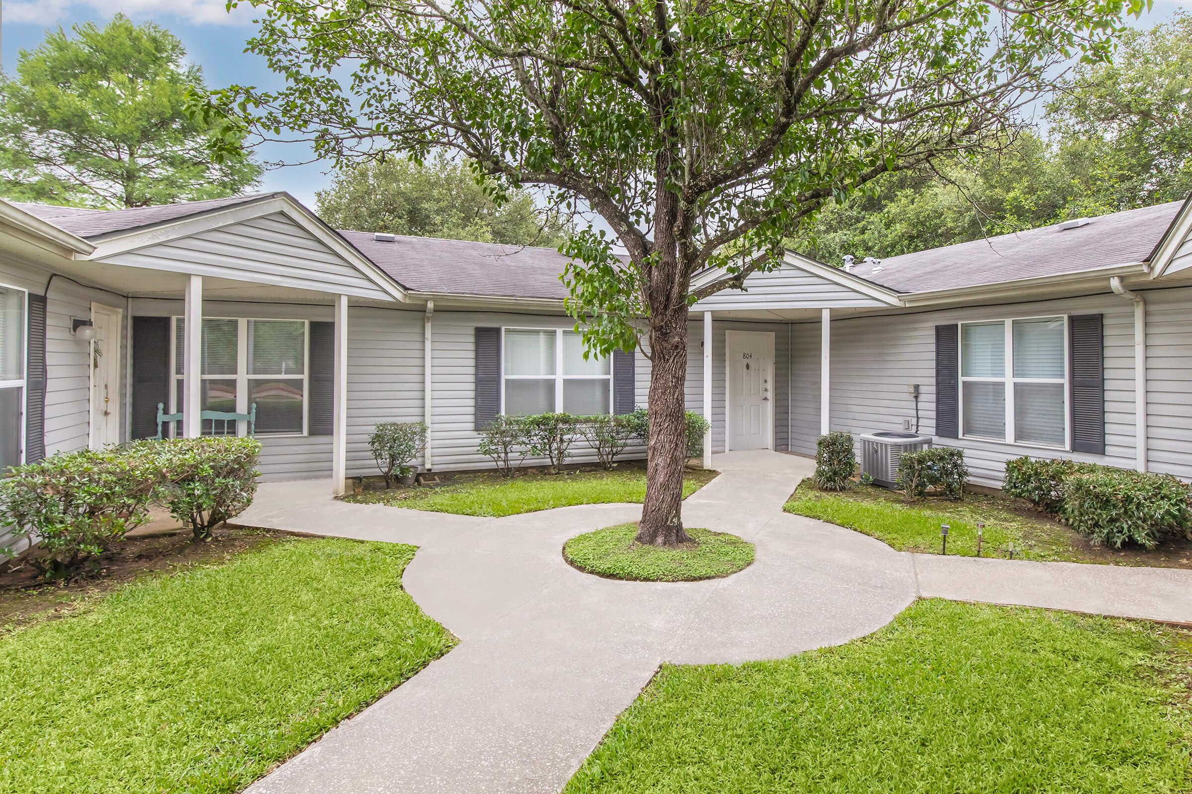 A landscaped courtyard featuring a central tree surrounded by a concrete pathway. The pathway leads to two single-story residential buildings, each with multiple windows and shutters. Lush green grass and small shrubs enhance the inviting atmosphere of the area on a sunny day.