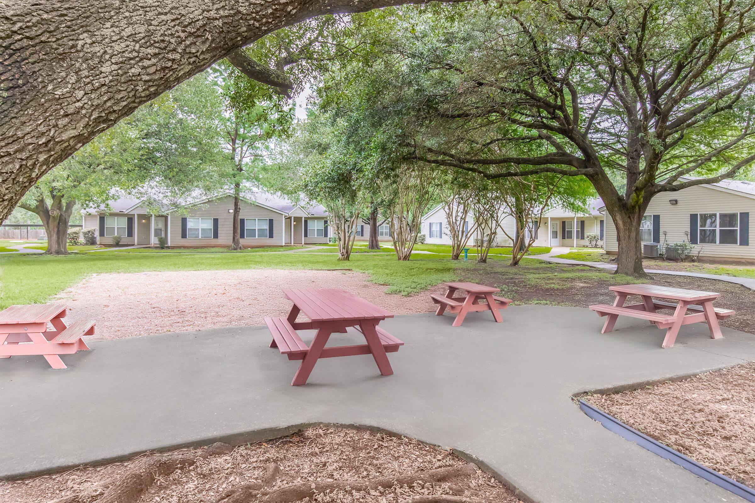 A tranquil outdoor space featuring several red picnic tables on a patio area, surrounded by lush greenery and trees. In the background, there are buildings with a simple, residential design, set amidst a grassy landscape. The scene invites relaxation and social gatherings.
