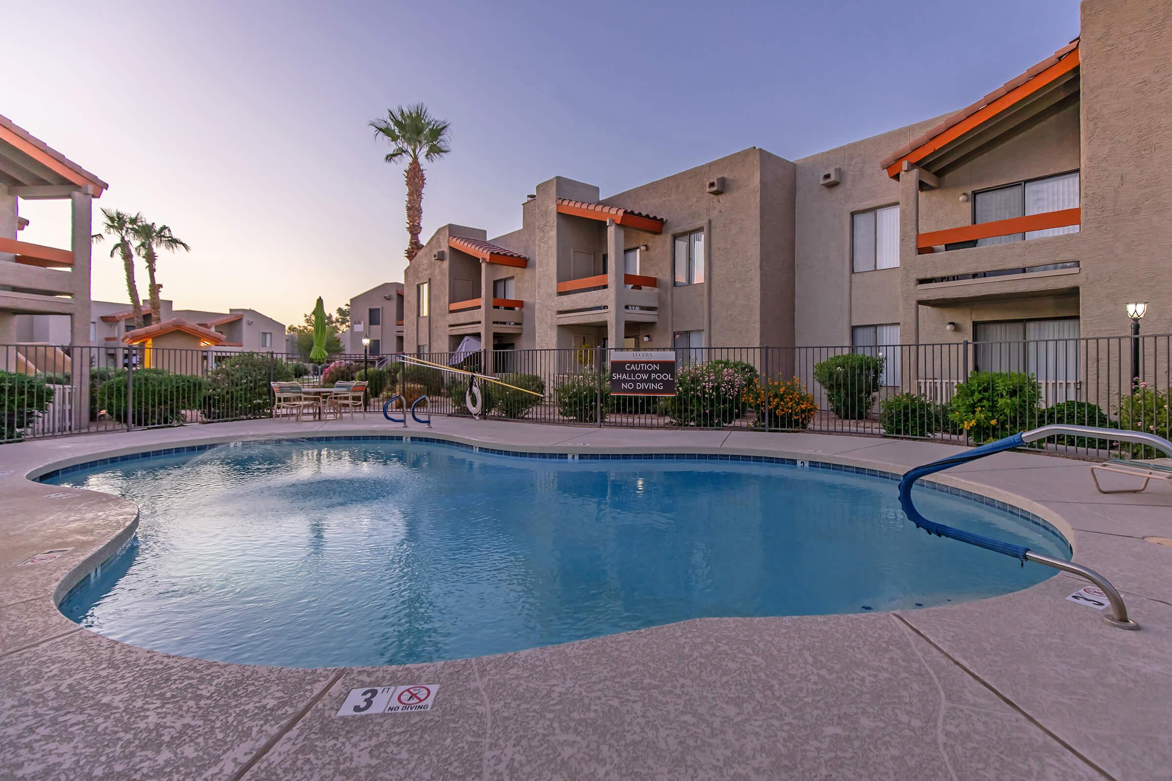 A serene swimming pool surrounded by lush greenery and palm trees, with lounge chairs nearby. The backdrop features modern apartment buildings with orange accents, illuminated by soft evening light, creating a relaxing atmosphere. A safety sign is visible near the pool area.