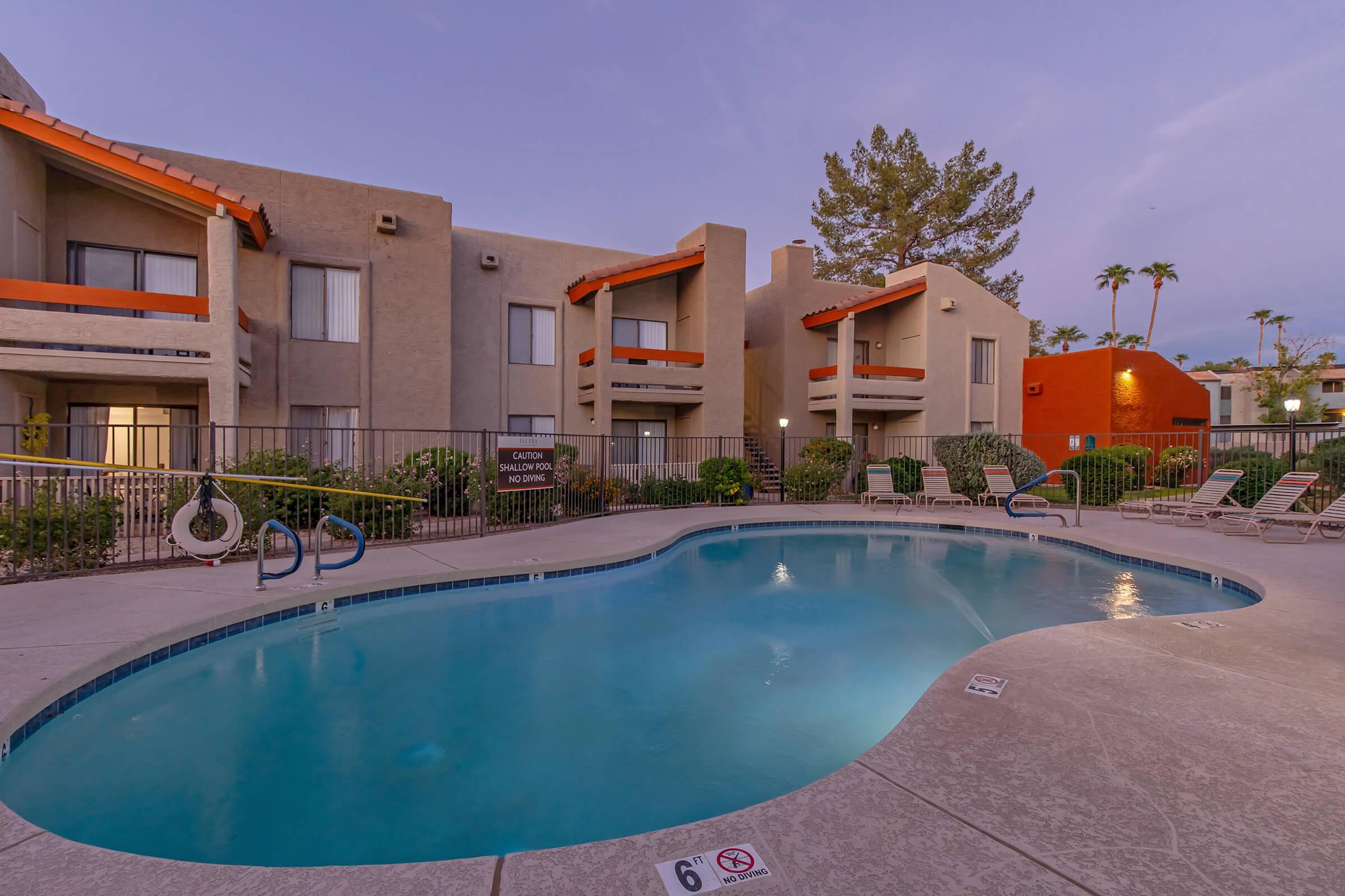 A tranquil residential pool area featuring a curved swimming pool surrounded by lounge chairs. The backdrop includes modern apartment buildings with balconies and palm trees, set against a twilight sky. A sign near the pool indicates it is for residents only.