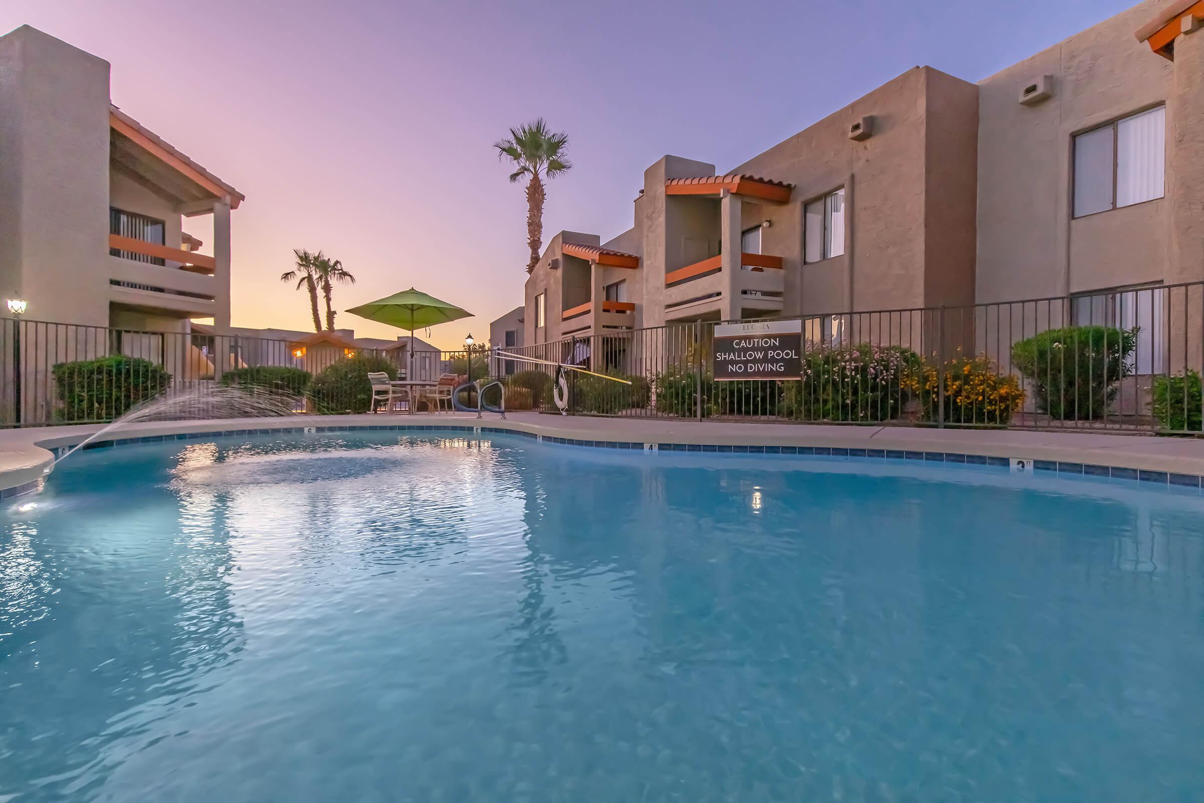 A tranquil swimming pool surrounded by greenery and palm trees, with residential buildings in the background. The sky is painted in soft hues of purple and pink during sunset. An umbrella provides shade by the poolside, and a sign indicates it's a shallow pool with a caution notice.