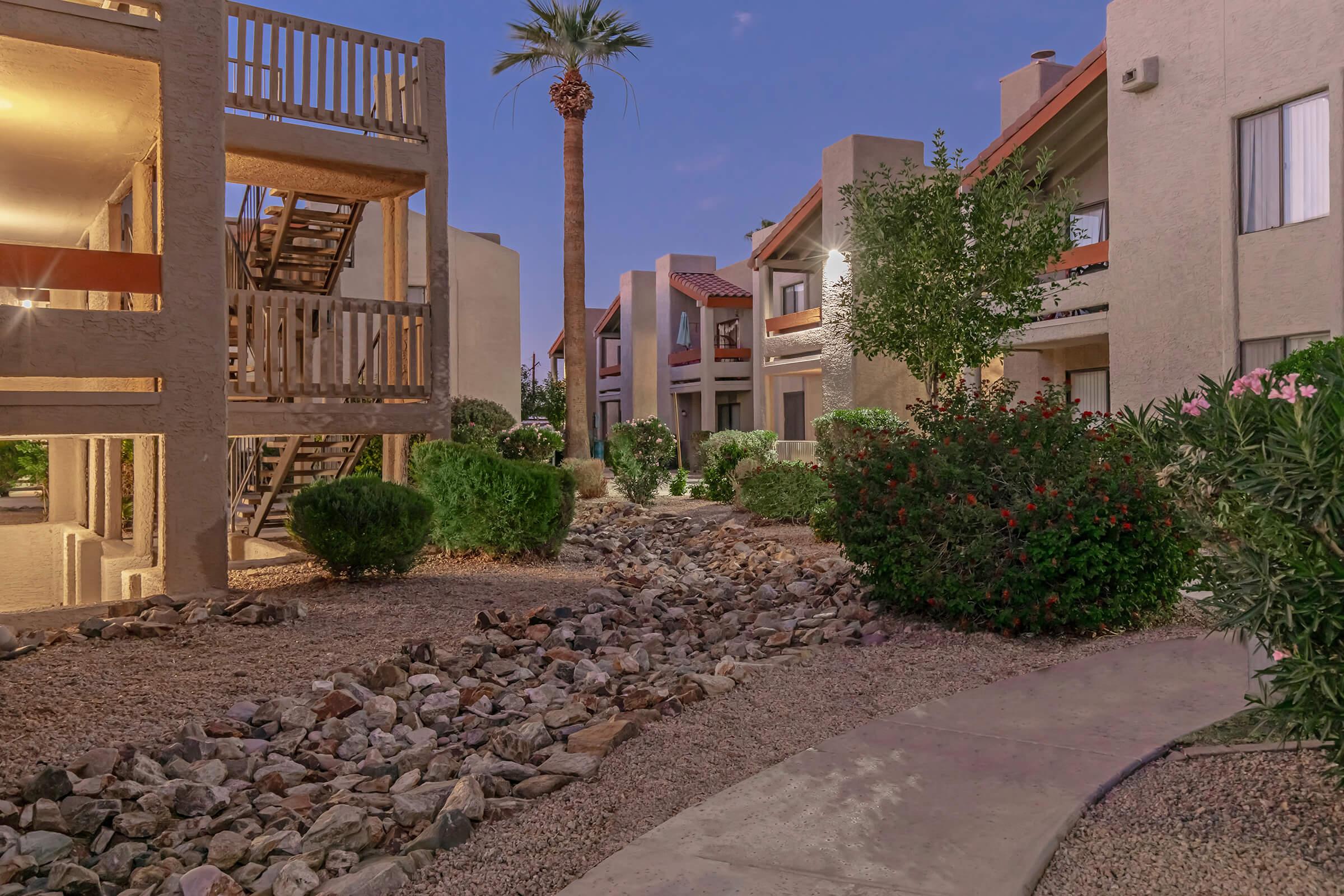 View of a landscaped apartment complex at dusk, featuring pathways, rock gardens, and lush greenery. The buildings have a mix of beige and red accents, with a palm tree visible in the background and outdoor staircases leading to upper units.