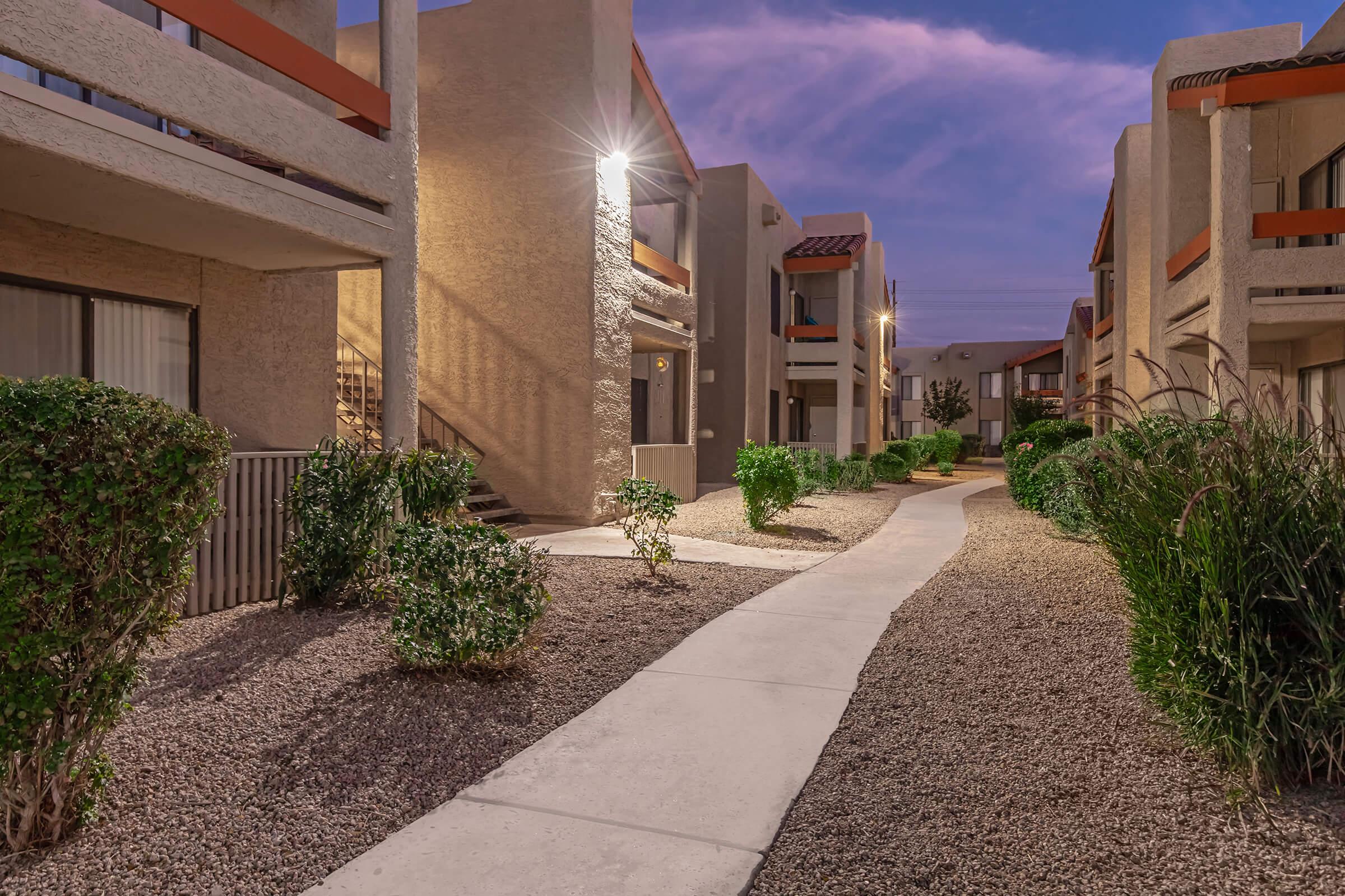 A tranquil pathway winding between two-story apartment buildings at dusk, with landscaped greenery, pebbled ground, and soft exterior lighting. The sky features muted shades of purple and blue, creating a serene atmosphere.