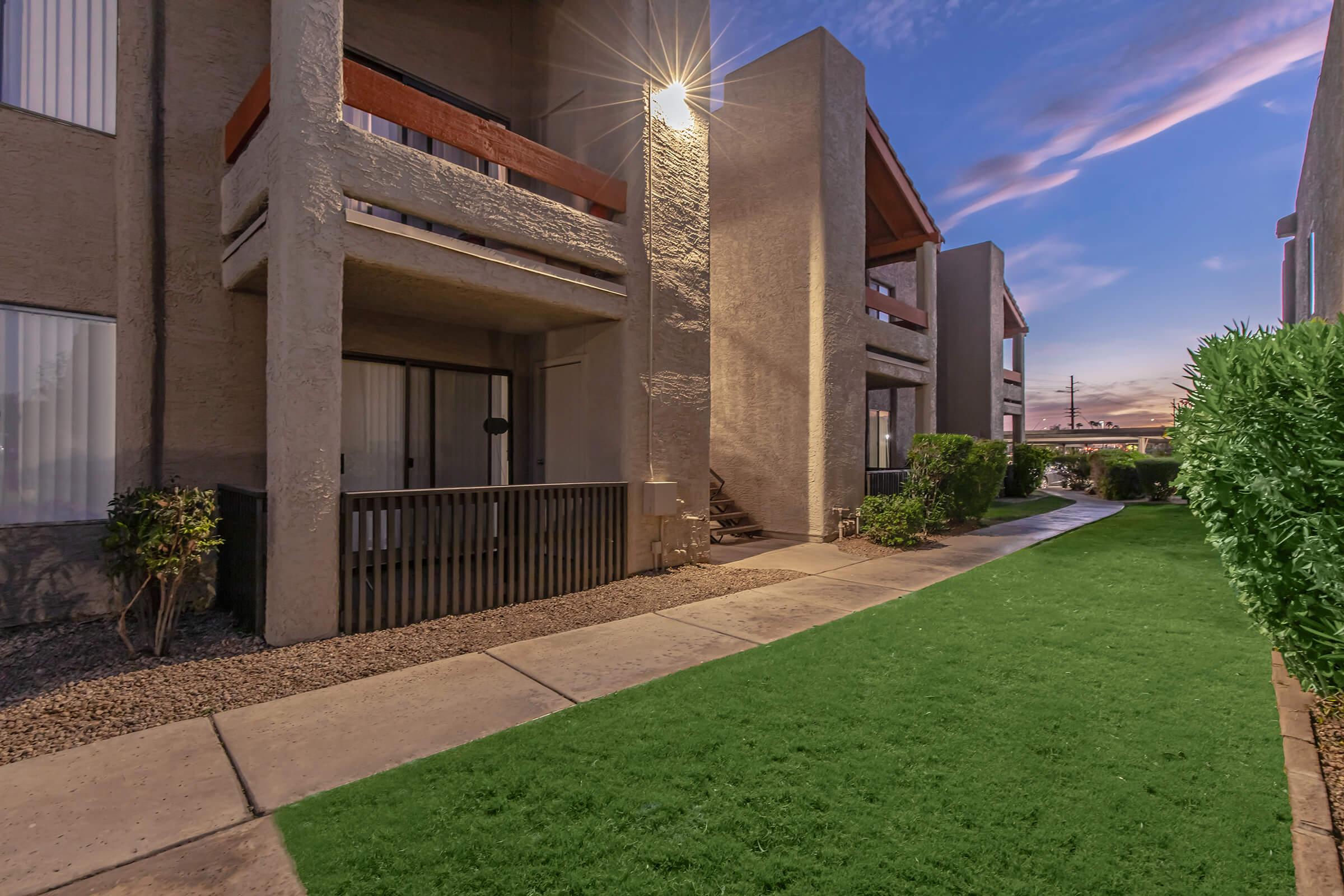 Sidewalk leading through a landscaped area with green grass and shrubs, flanked by two multi-story apartment buildings. The scene is illuminated by a light from one of the balconies, with a clear blue sky in the background at dusk.