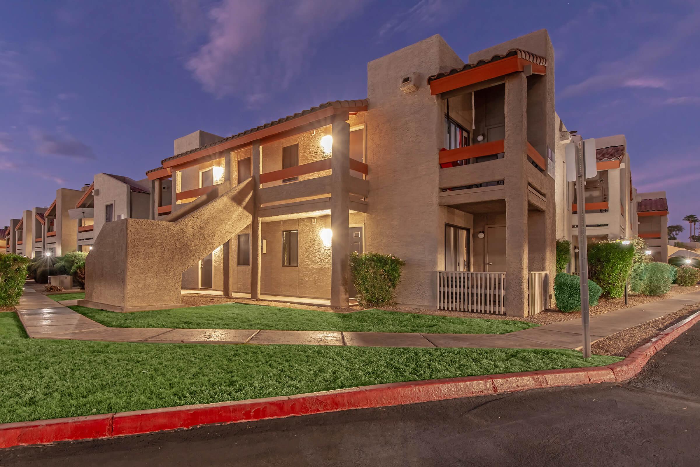 A modern apartment building with a stucco exterior, featuring balconies and an exterior staircase. The scene is set during dusk, with lights on inside the units. Lush green grass and well-maintained landscaping surround the building, adding to the inviting atmosphere.