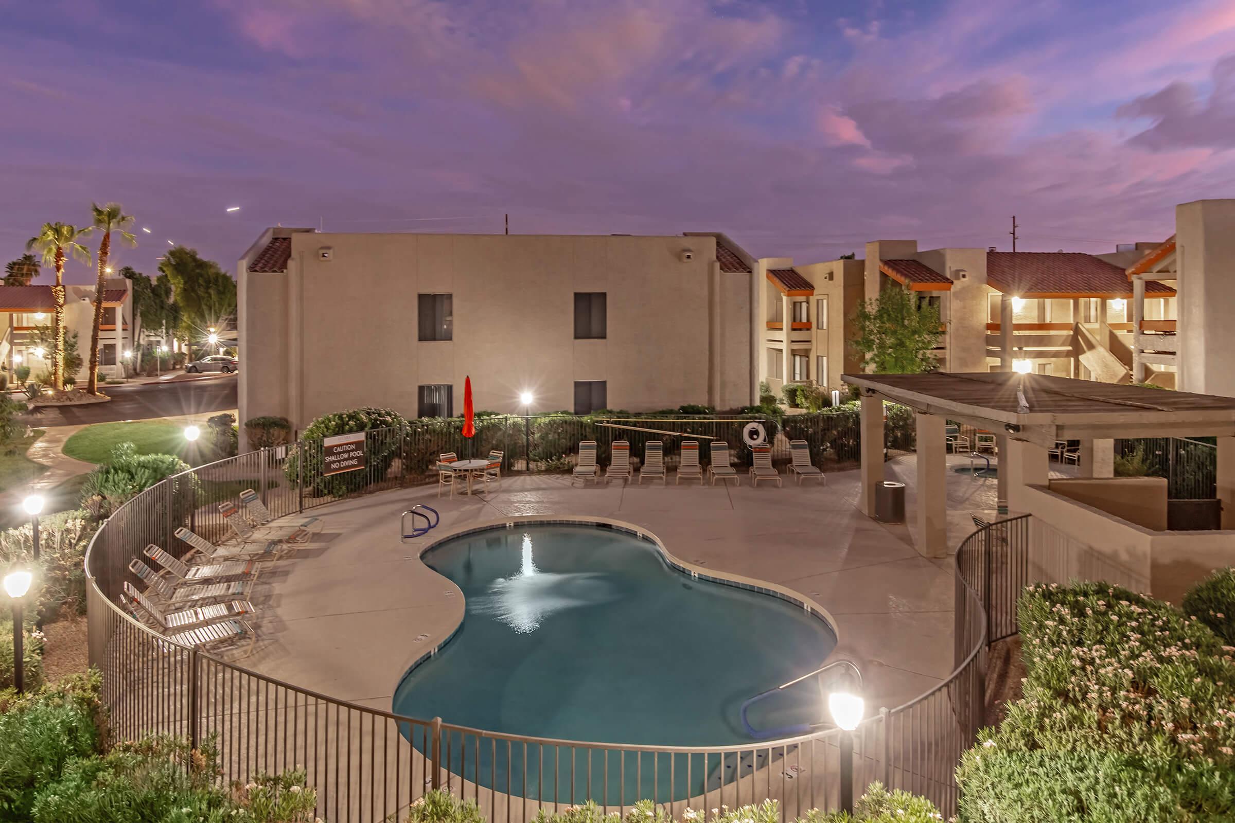 A serene evening view of a swimming pool surrounded by lush greenery and lounge chairs, with two apartment buildings in the background. The scene is illuminated by soft outdoor lighting under a colorful sky during twilight.