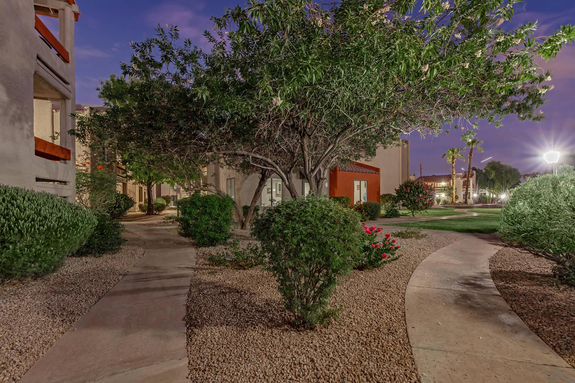 A serene outdoor pathway winding through landscaped gardens with shrubs and trees. The scene is illuminated by soft lighting in the evening, showcasing a peaceful residential area with nearby buildings and green spaces.