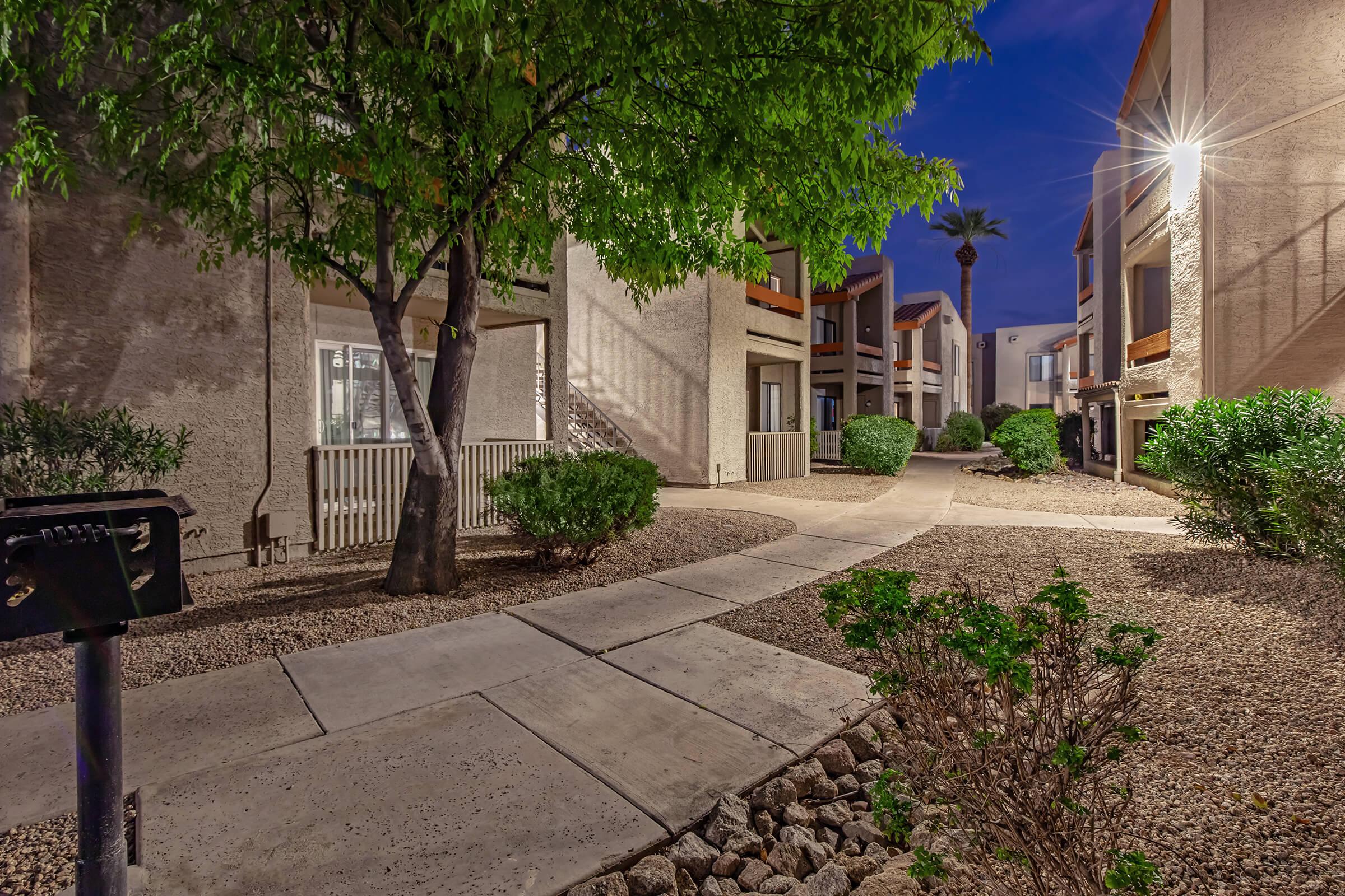 A walkway winding through a well-maintained courtyard of a residential complex, featuring light-colored buildings, sparse landscaping with bushes, and a tree. The scene is illuminated by artificial lighting at dusk, creating a serene atmosphere.