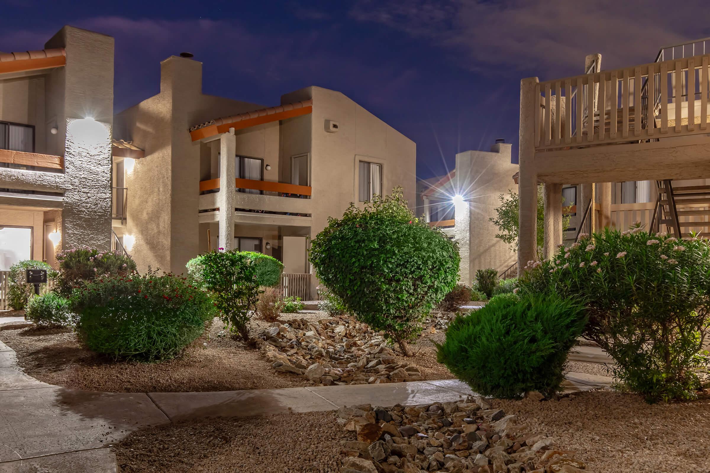 A landscaped courtyard area at night featuring several buildings with illuminated windows. Lush greenery, neatly trimmed bushes, and a gravel pathway create a serene atmosphere. Soft lighting highlights the architectural details of the surrounding structures.
