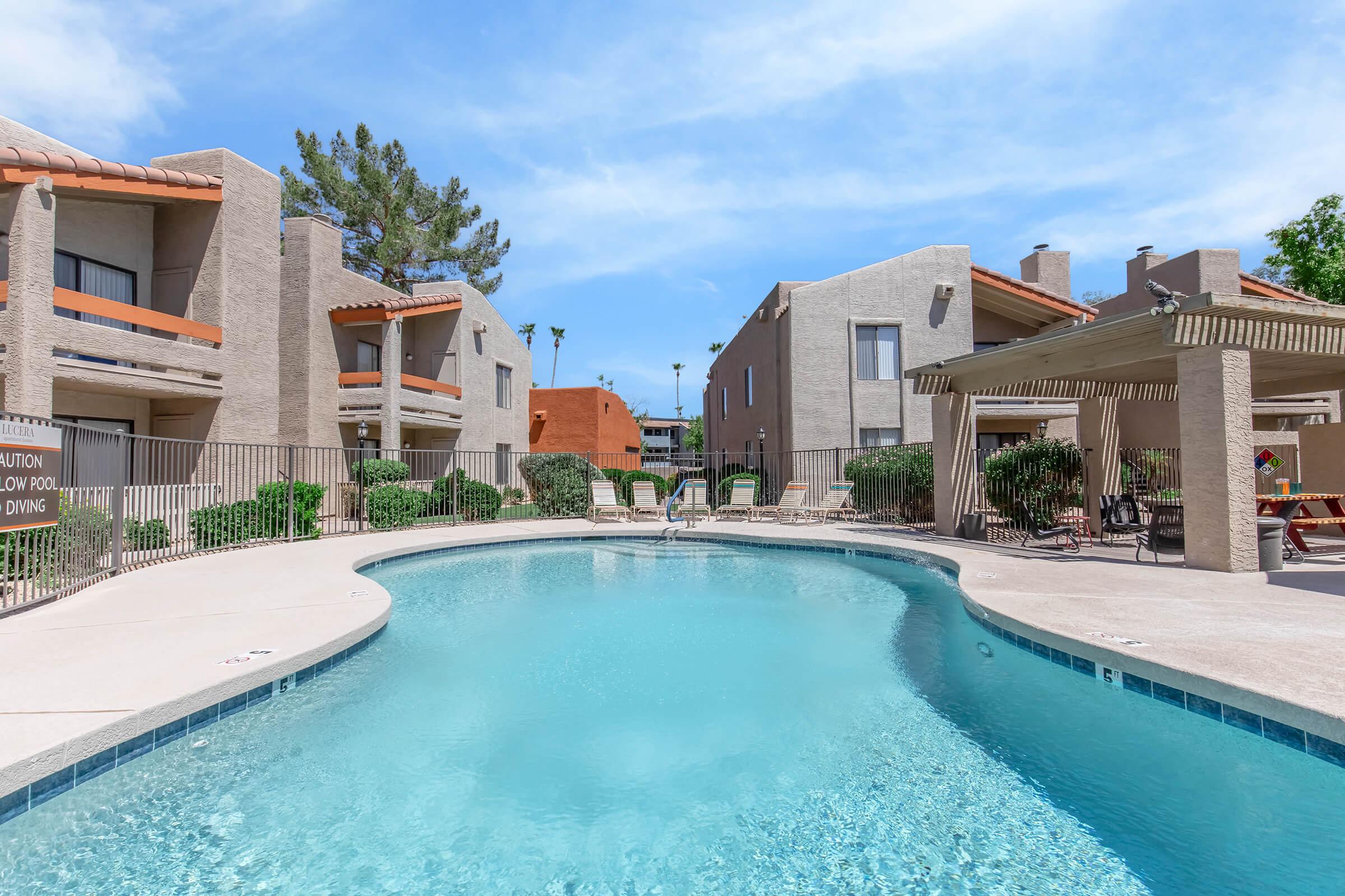 A tranquil apartment complex featuring a clear swimming pool surrounded by lounge chairs, palm trees, and landscaped greenery. The buildings are two stories high, with balconies overlooking the pool area, under a bright blue sky with a few clouds.