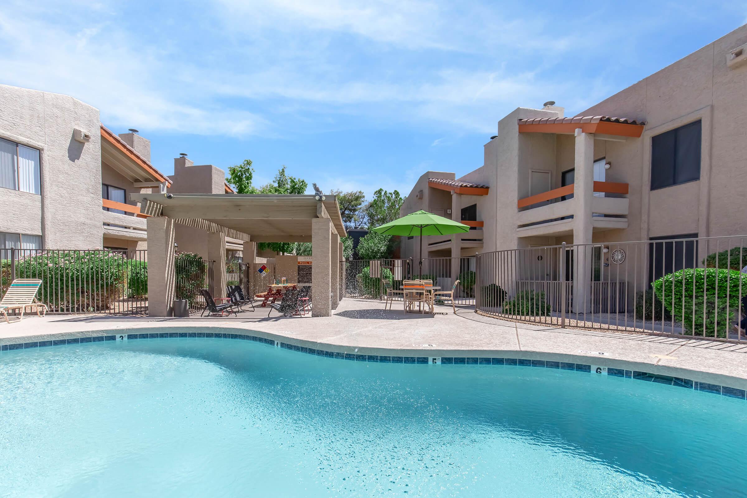 A clear blue swimming pool surrounded by a landscaped area with lounge chairs, a shaded pergola, and an umbrella. Nearby, two multi-story residential buildings are visible, with balconies and greenery around the pool area under a bright blue sky.