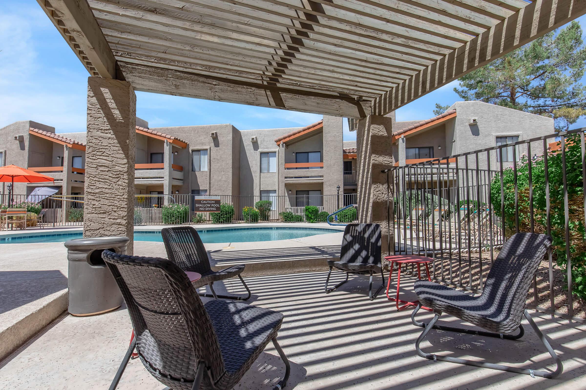 A shaded outdoor seating area with black wicker chairs and a small red table, overlooking a swimming pool. In the background, there are apartment buildings with orange roofs and greenery. The setting is bright and sunny, typical of a residential complex.