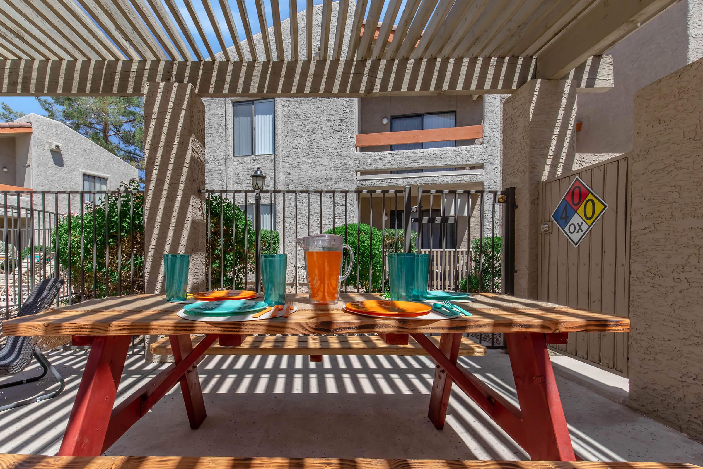 Outdoor dining setup on a shaded picnic table with colorful plates and glasses. A large pitcher sits in the center, and the background features a gated area with greenery and a sign. The scene is bright with sunlight filtering through the slatted pergola above.
