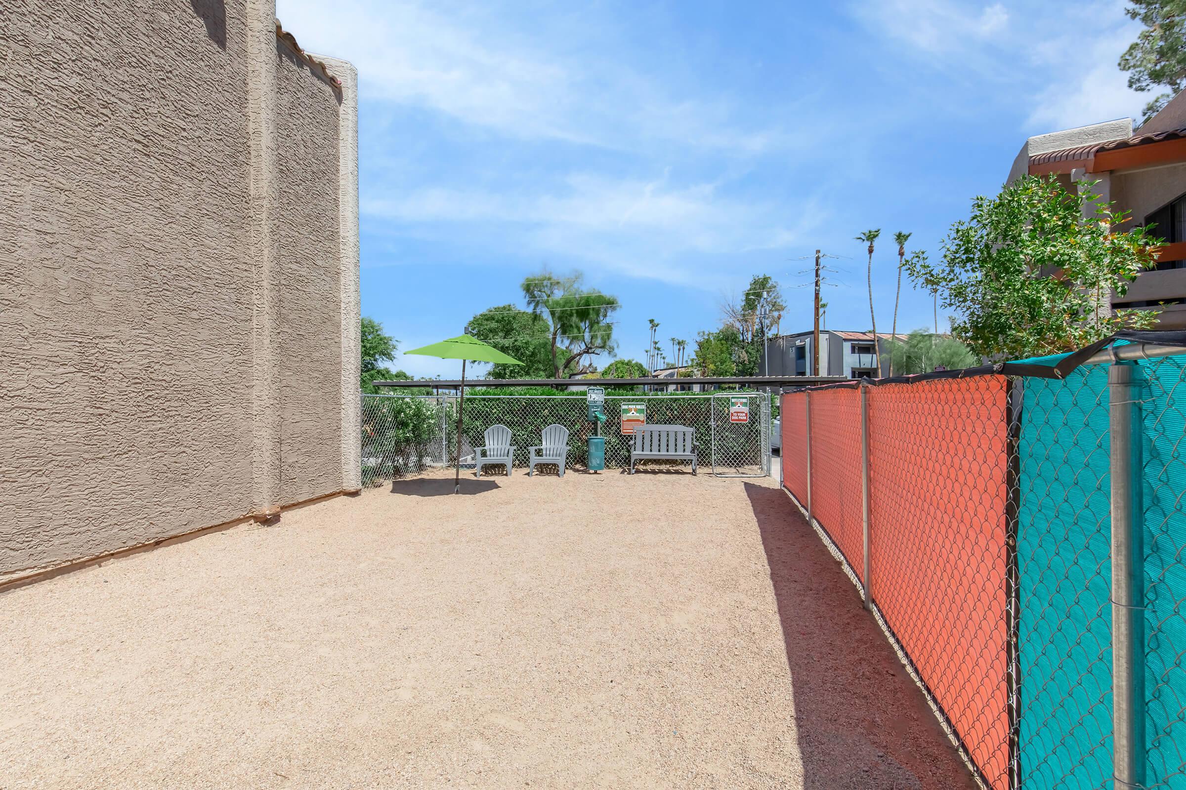 A fenced outdoor area with gravel ground and a few chairs, surrounded by buildings and greenery. There are trees and a gazebo in the background, creating a tranquil setting suitable for relaxation. The sky is clear with some clouds.