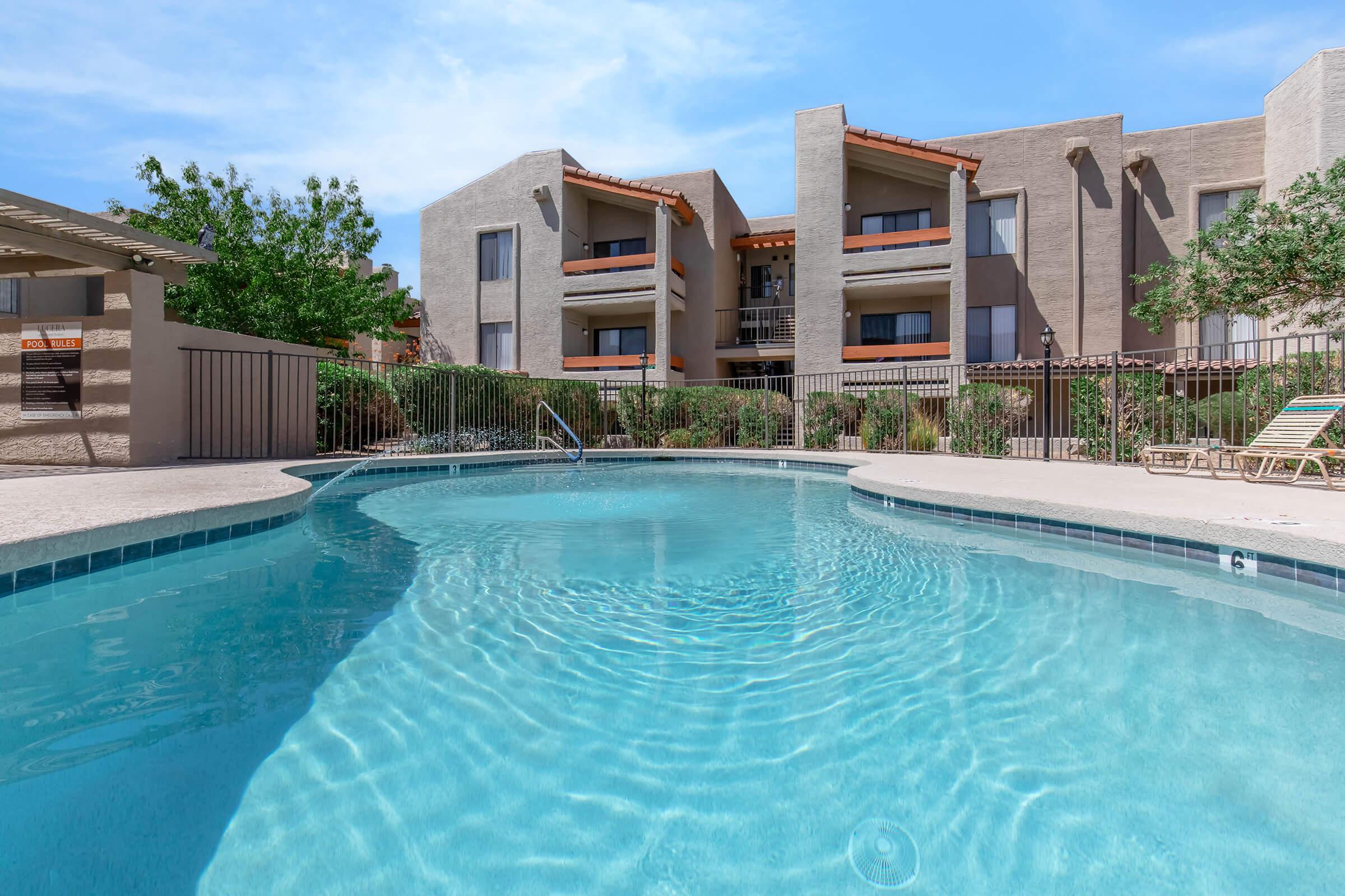A clear swimming pool with gentle ripples in the water, surrounded by a fenced area. In the background, there are multi-story apartment buildings with orange tile roofs and lush greenery. A sunny sky with a few clouds completes the scene, creating a relaxing outdoor atmosphere.