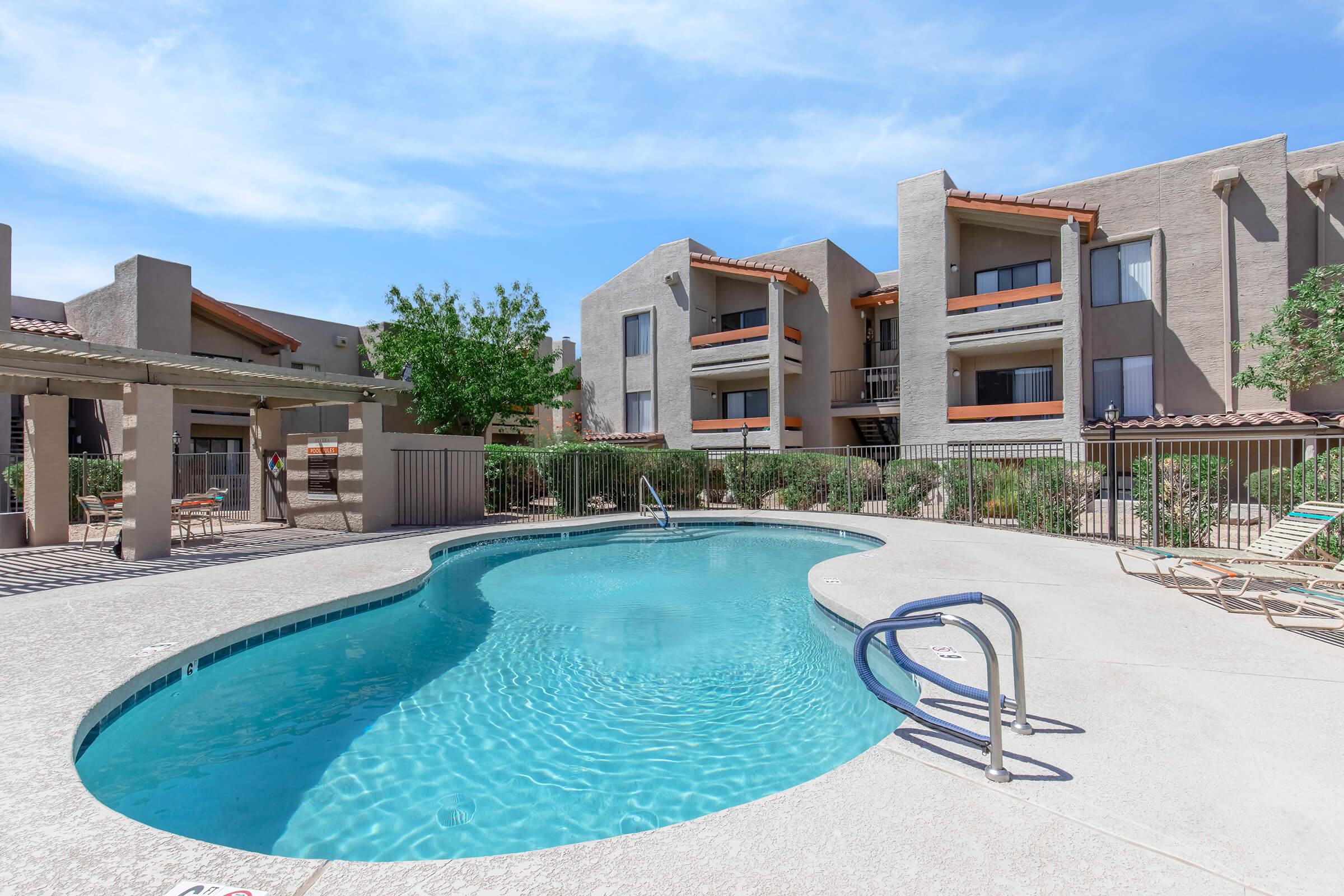 A clear blue swimming pool surrounded by a fenced area, with lounge chairs along the edge. In the background, there are modern apartment buildings with a desert landscape and bright blue sky above.