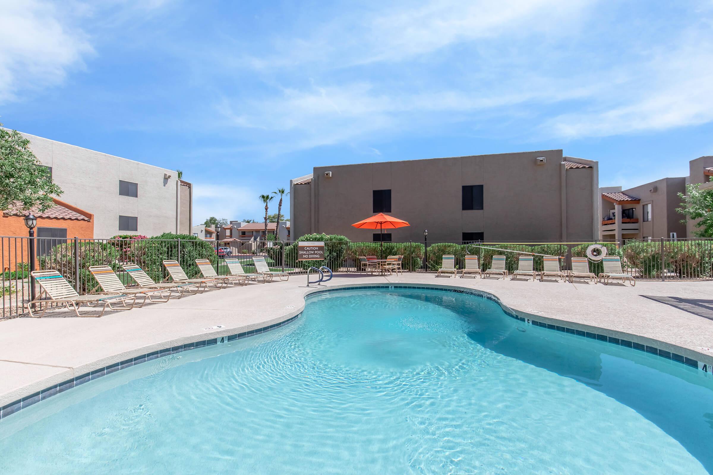 A serene outdoor swimming pool surrounded by lounge chairs and a sun umbrella, set against modern buildings and greenery under a clear blue sky.