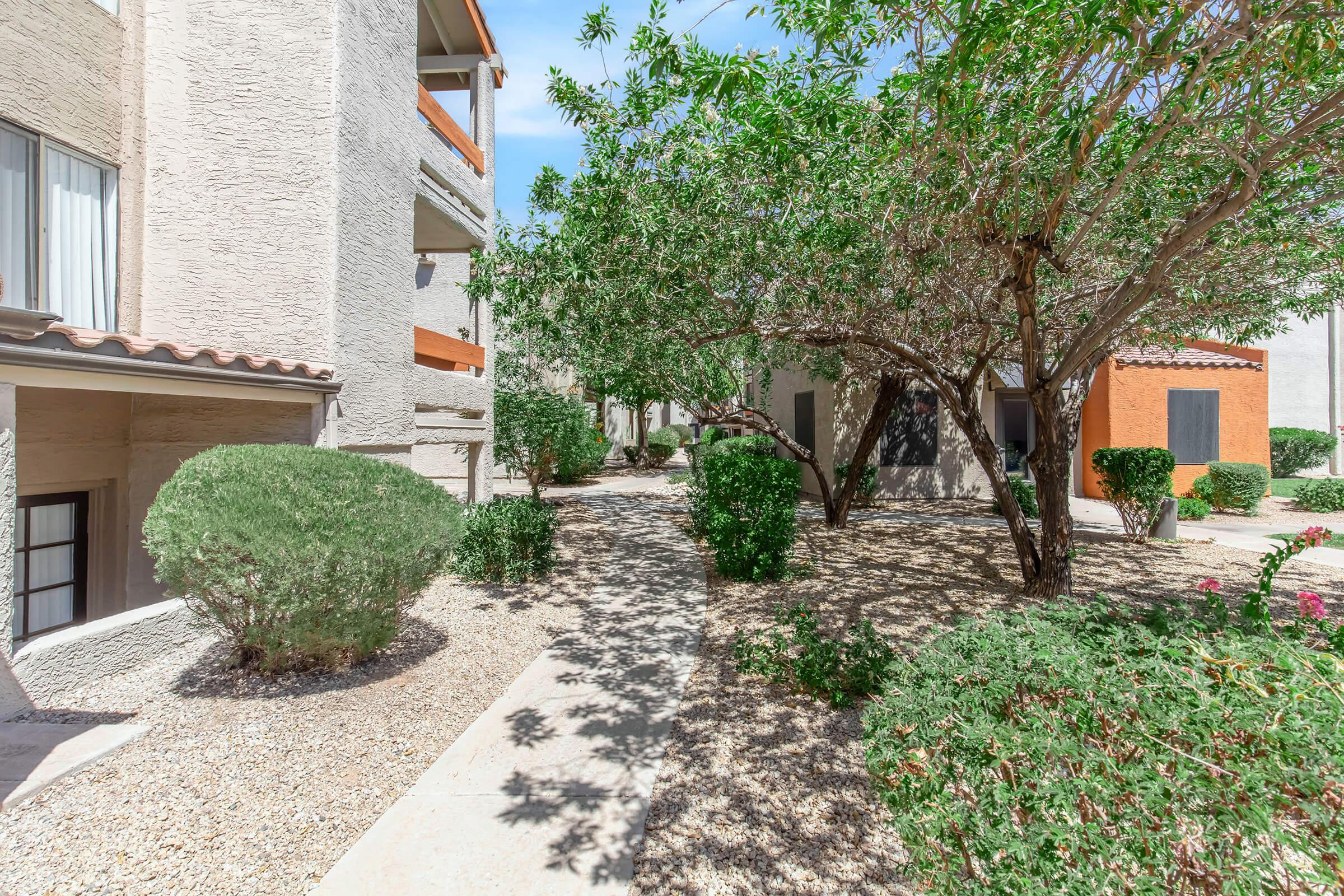 A landscaped pathway surrounded by greenery, featuring trees and shrubs, leading through an apartment complex. The buildings are light-colored with some vibrant orange accents in the background, set against a clear blue sky.