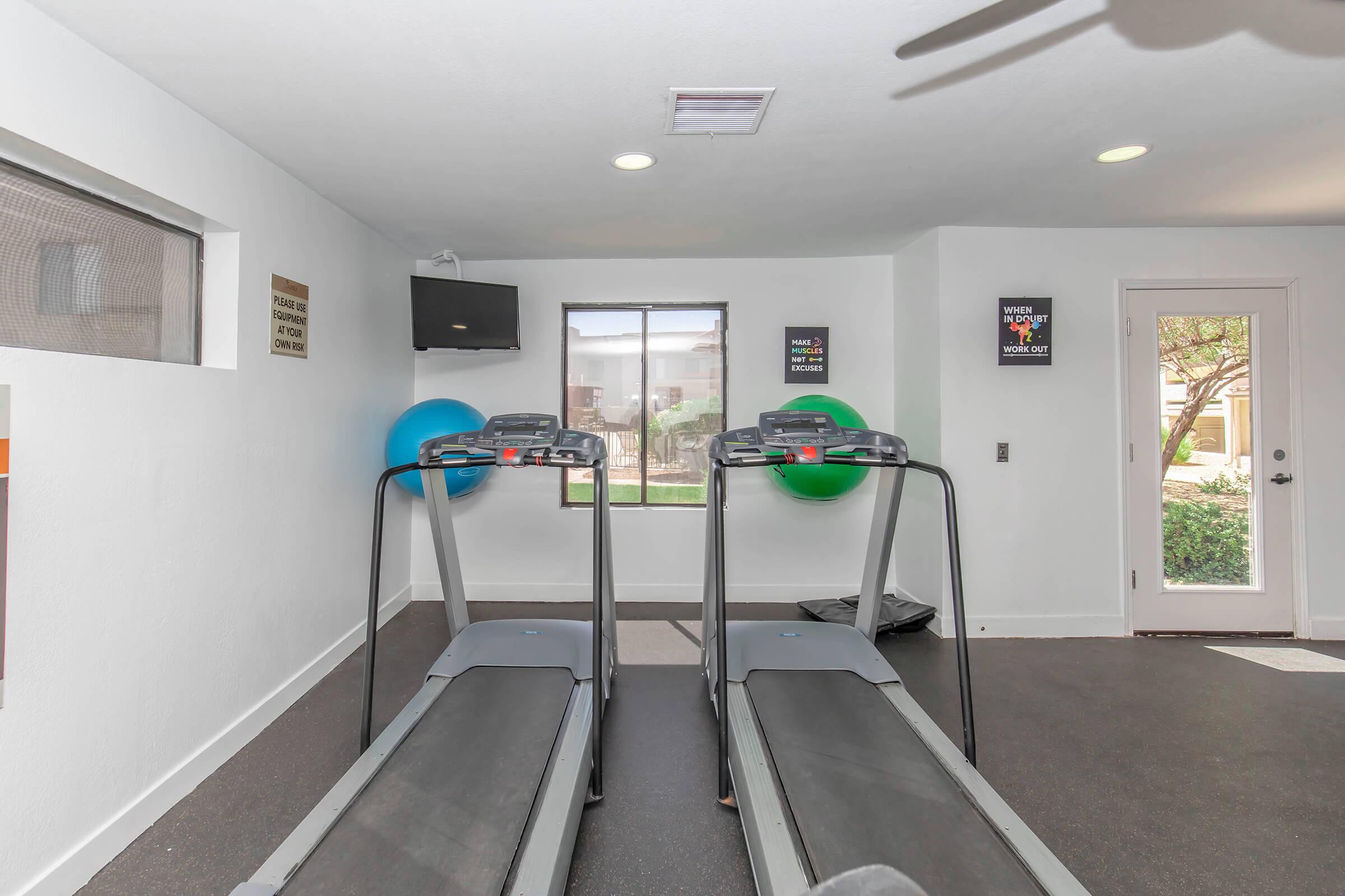 A fitness room featuring two treadmills, exercise balls, a television on the wall, and a window providing natural light. The space has a clean, modern design with motivational signs on the walls and a door leading outside.